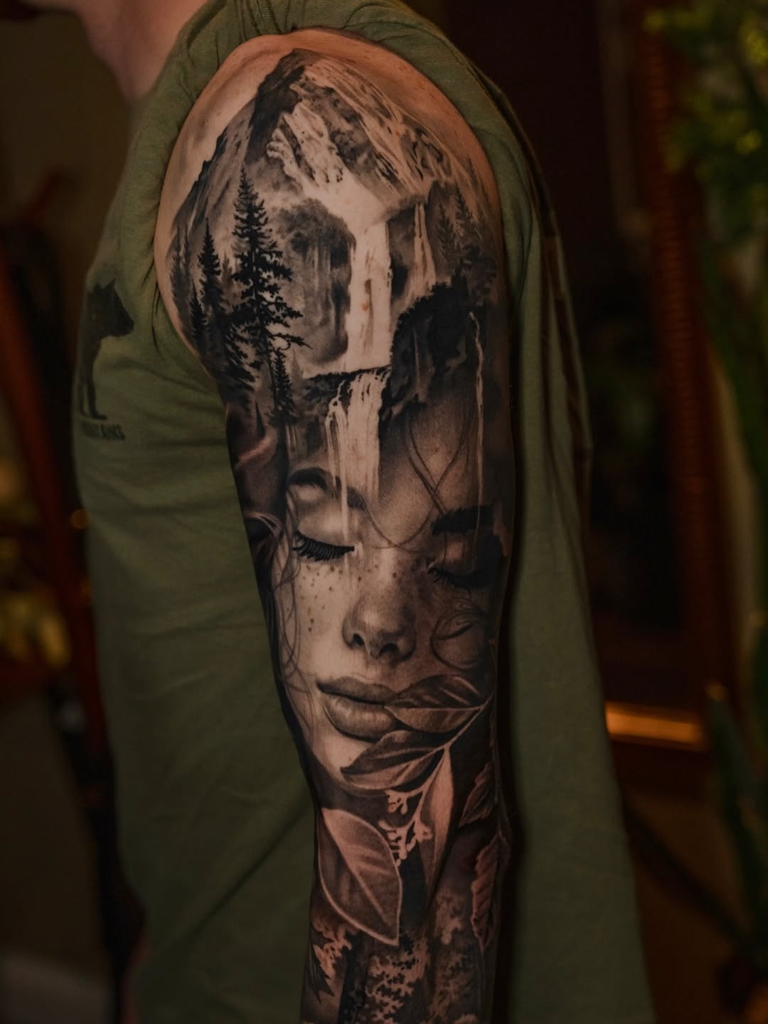 Woman face with mountain and waterfall surrealism tattoo on the upper arm by @kotytattoos