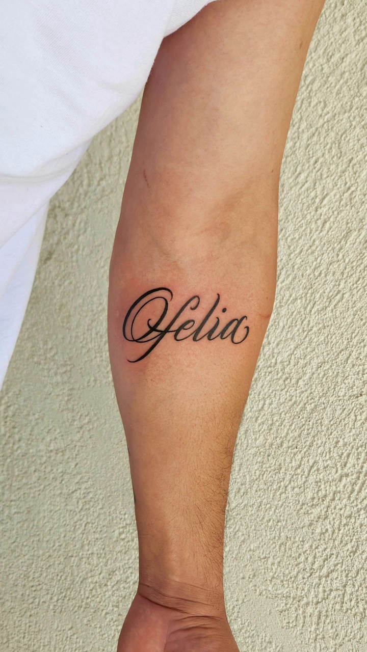 The name "Ofelia" in script lettering tattoo on the forearm by @youxg_guxxz