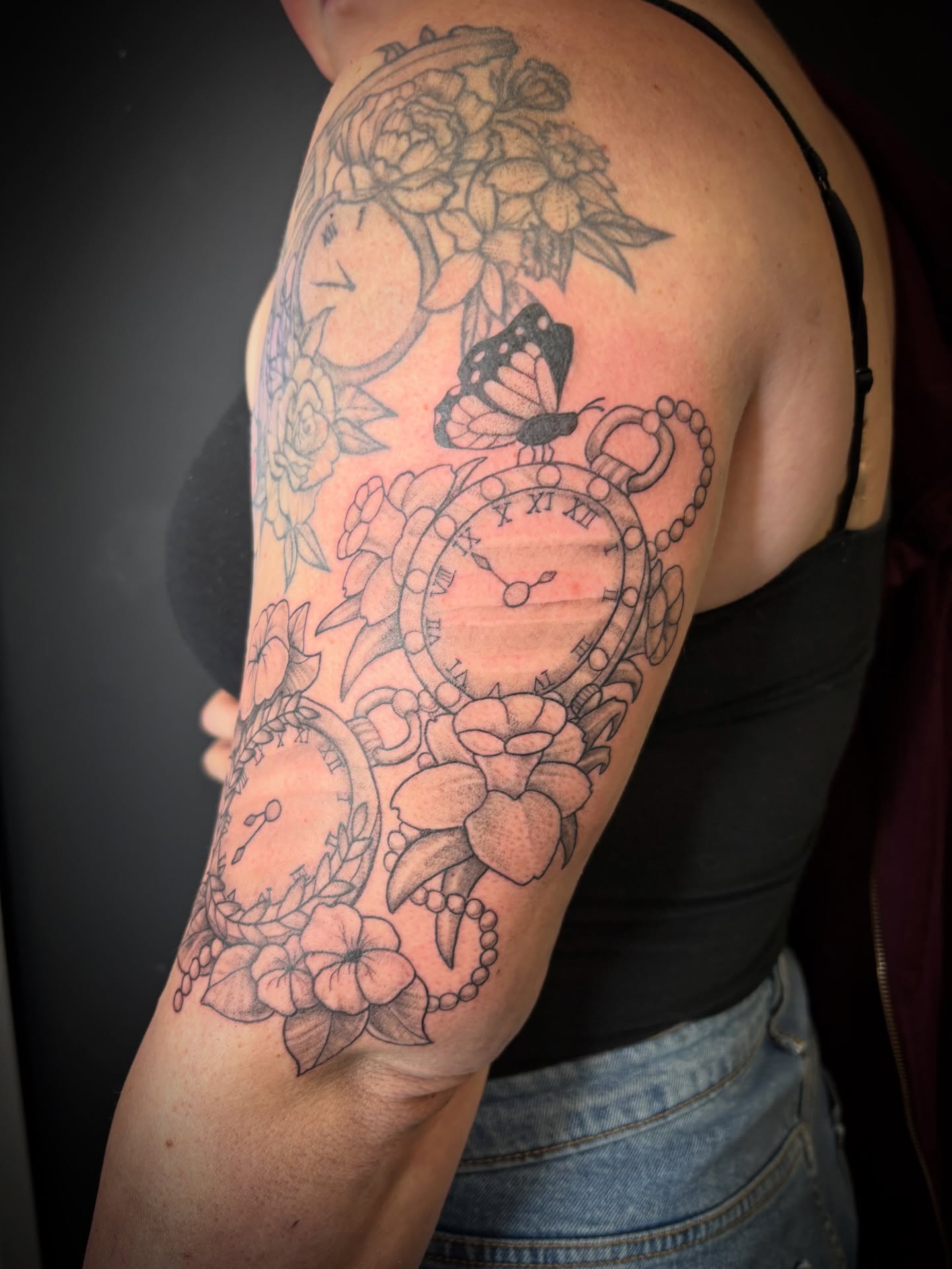 Clock and flowers black and grey tattoo on the upper arm by Noah Blum