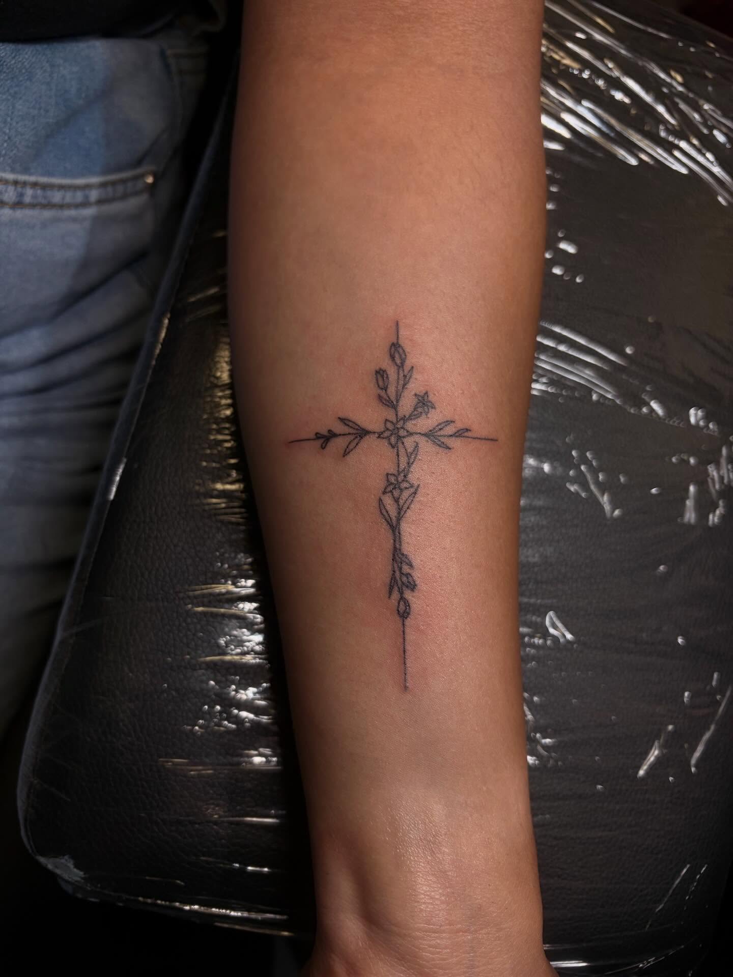 Cross with floral elements fine line tattoo on the forearm by Melissa