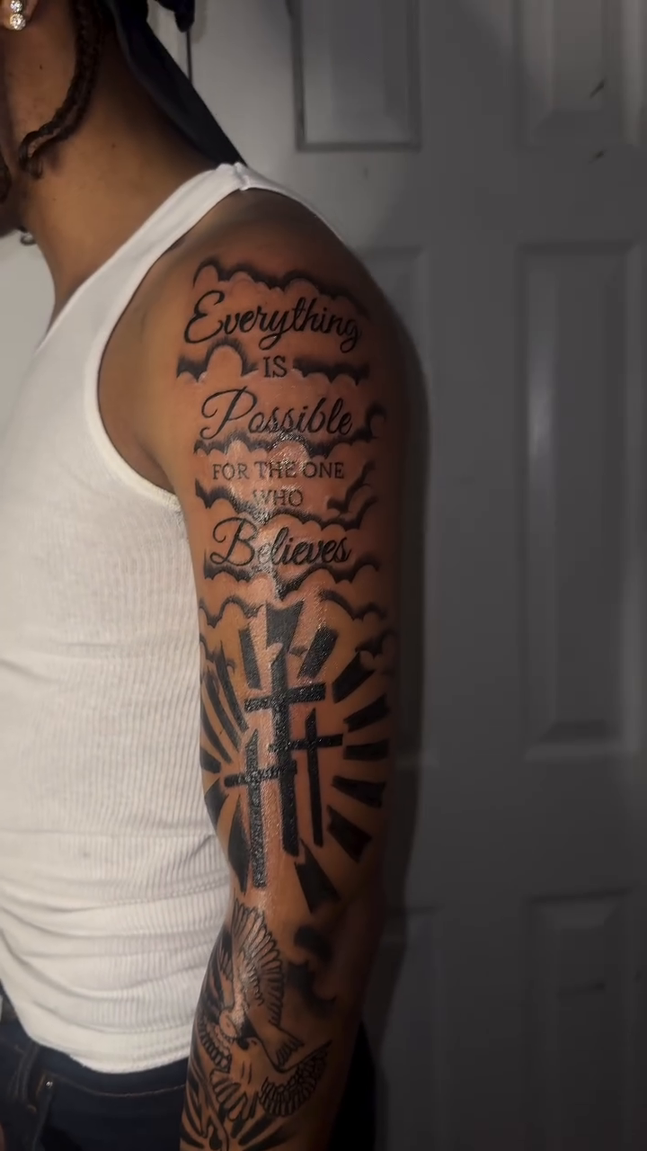 Cross with sunburst and lettering Chicano tattoo on the upper arm by Comet