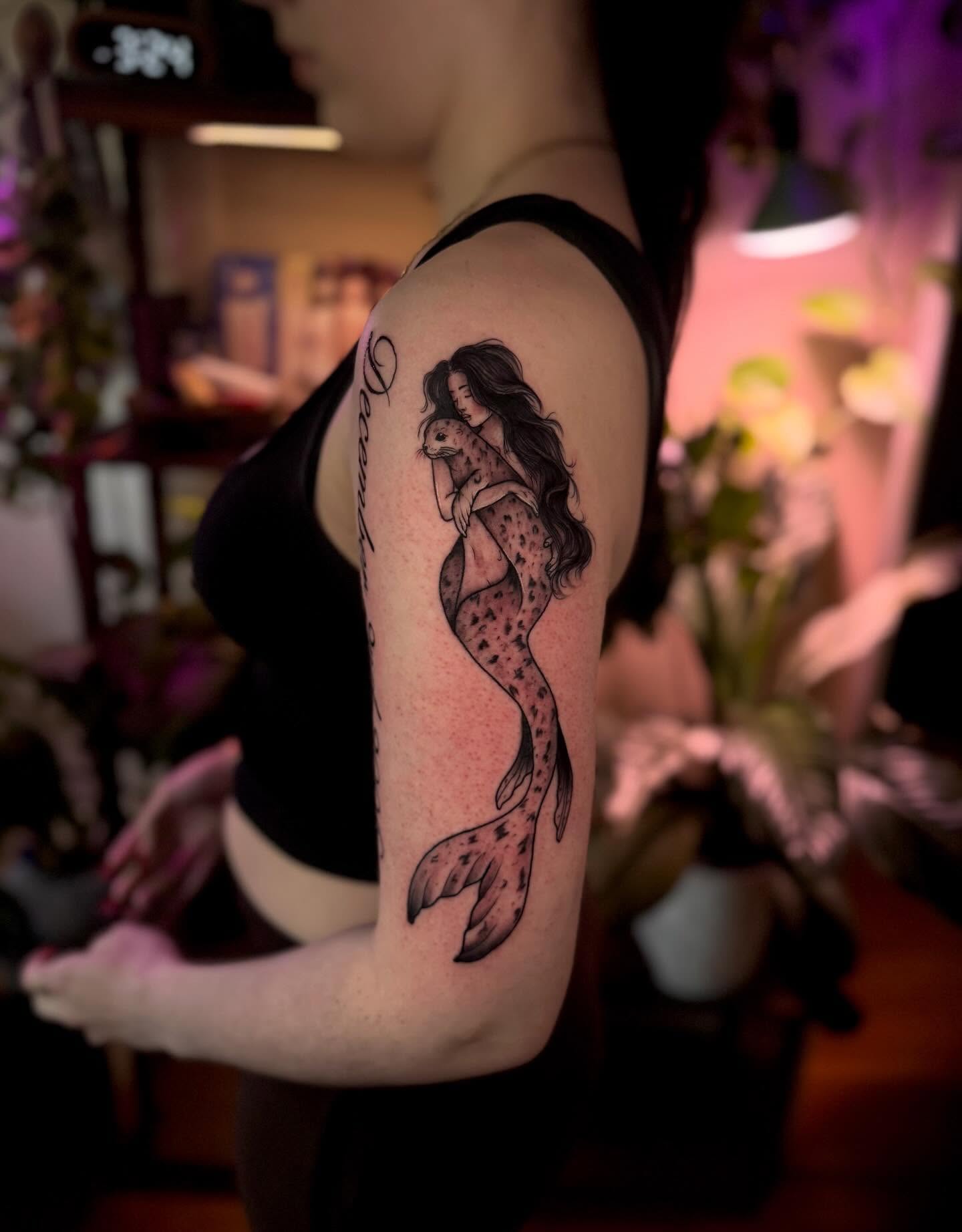 Mermaid holding seal black and grey tattoo on the upper arm by Angie Cotton