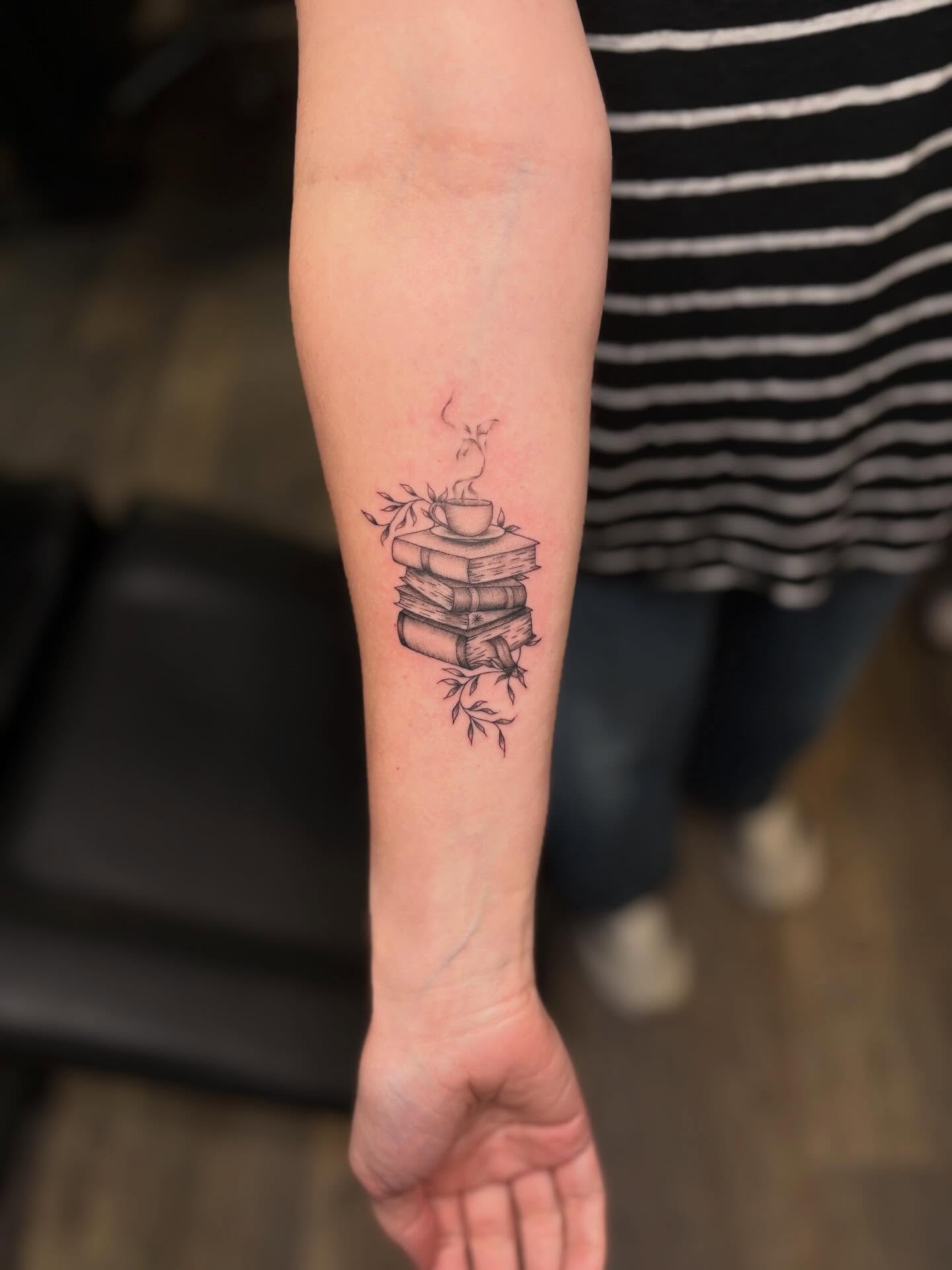 Stack of books with coffee cup black and grey tattoo on the forearm by Tessa Simmons - Tattoo Artist