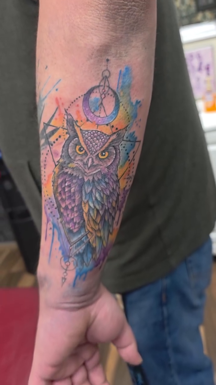 Owl with compass and watercolor splashes watercolor tattoo on the forearm by Bosco Layne