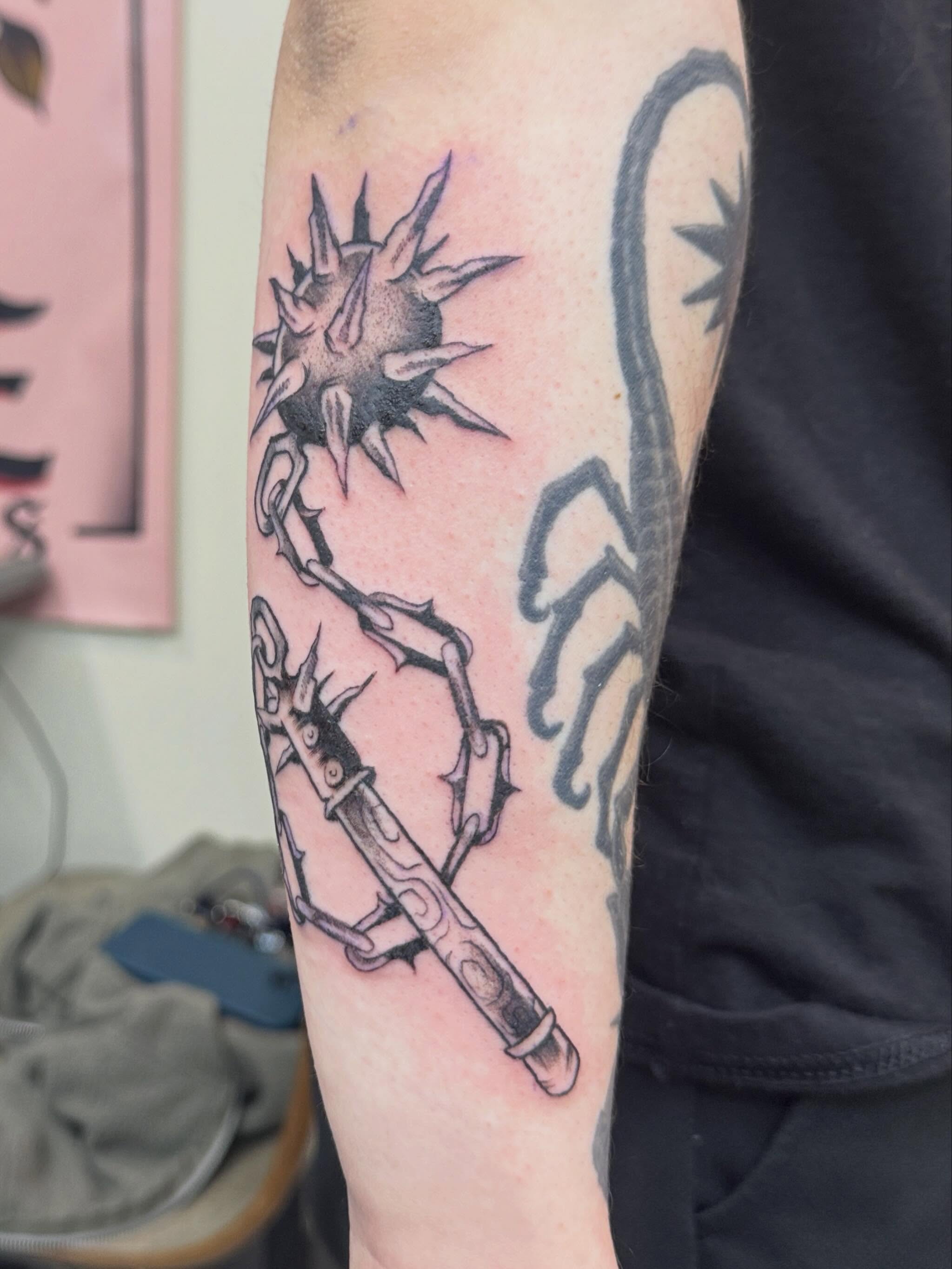 Gun and spiky object black and grey tattoo on the forearm by Rebecca Rose