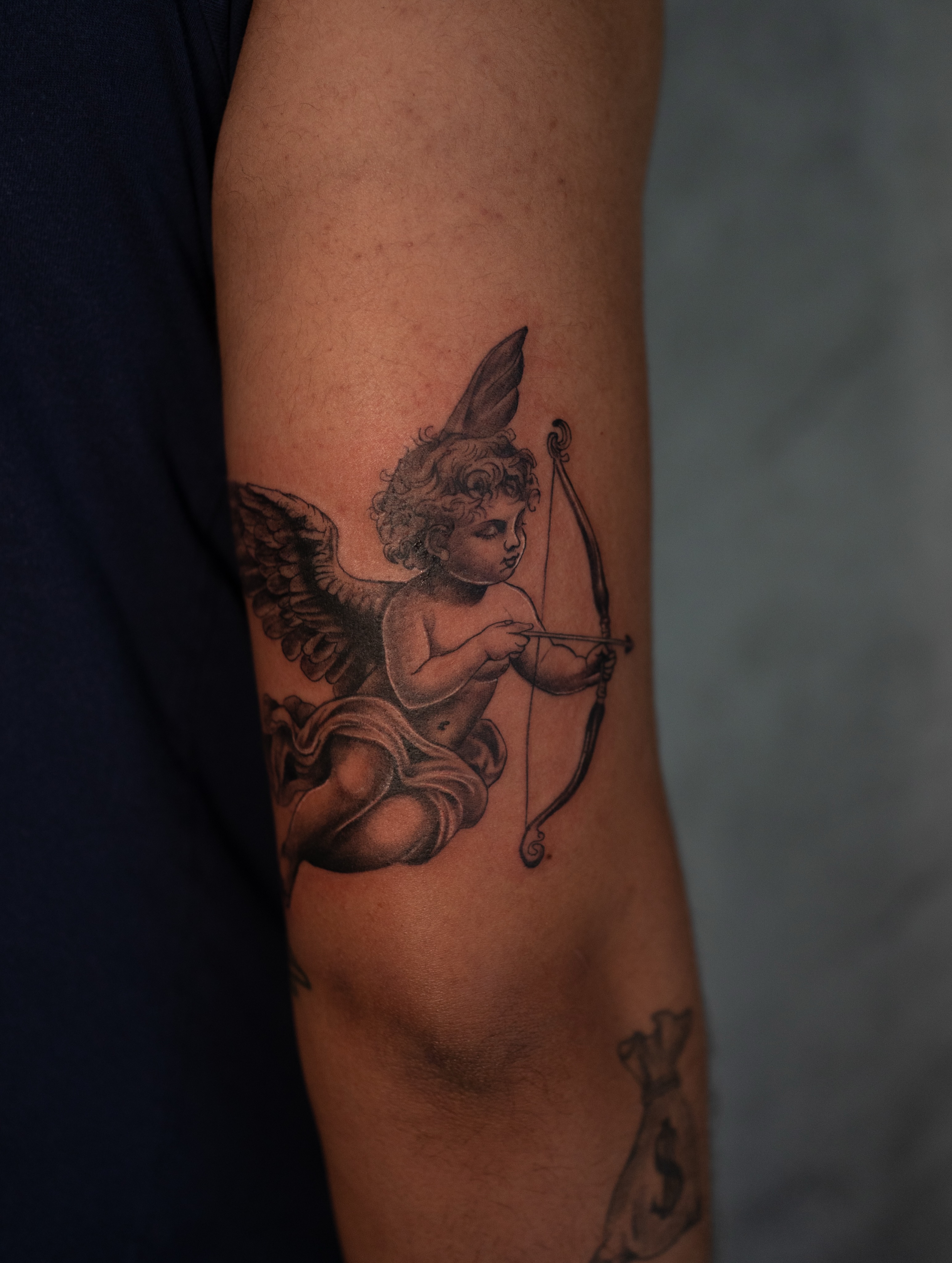 Cherub with bow black and grey tattoo on the forearm by Damon Valdez