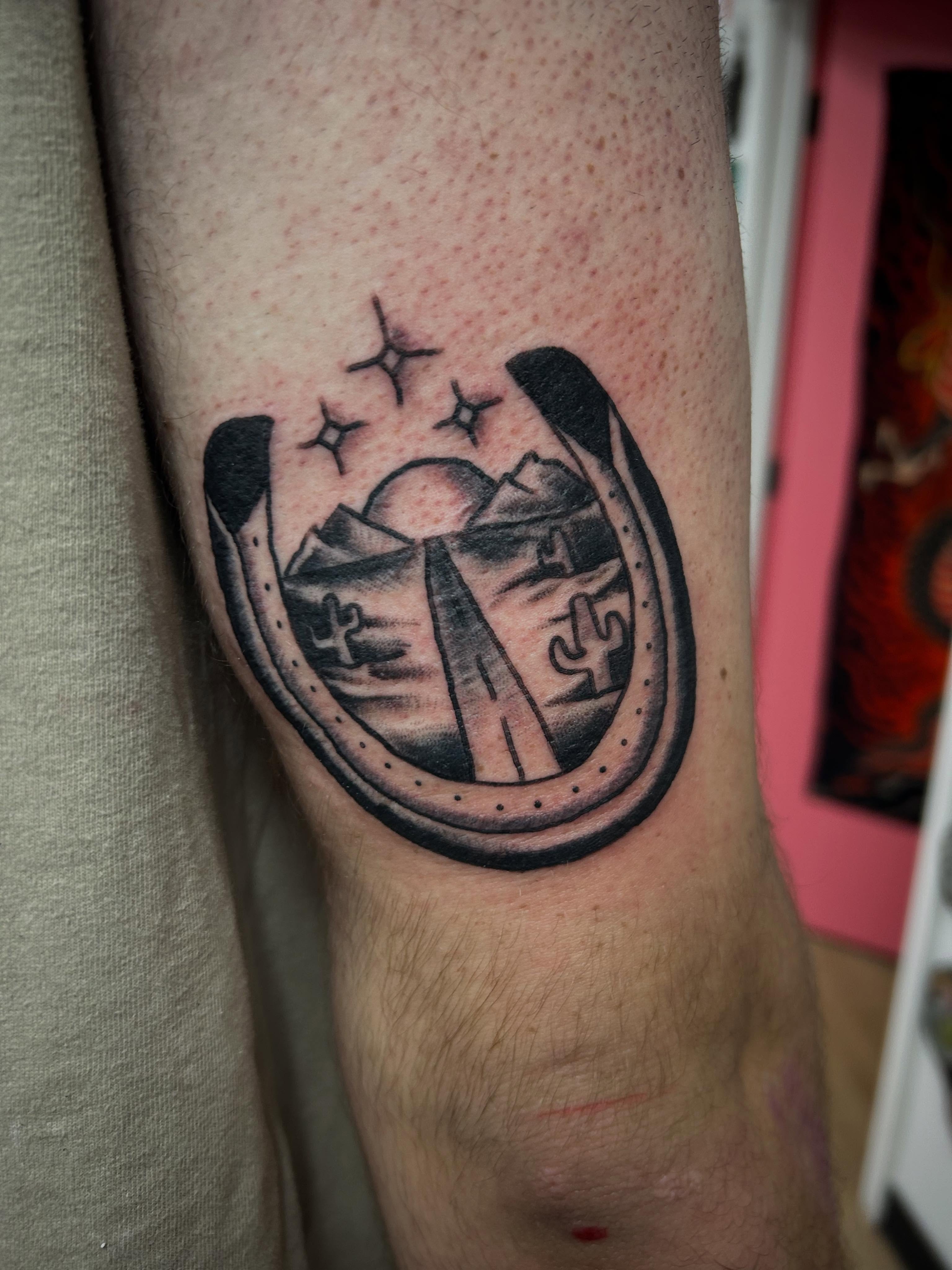 Horseshoe with desert landscape black and grey tattoo on the forearm by Stogie Tony
