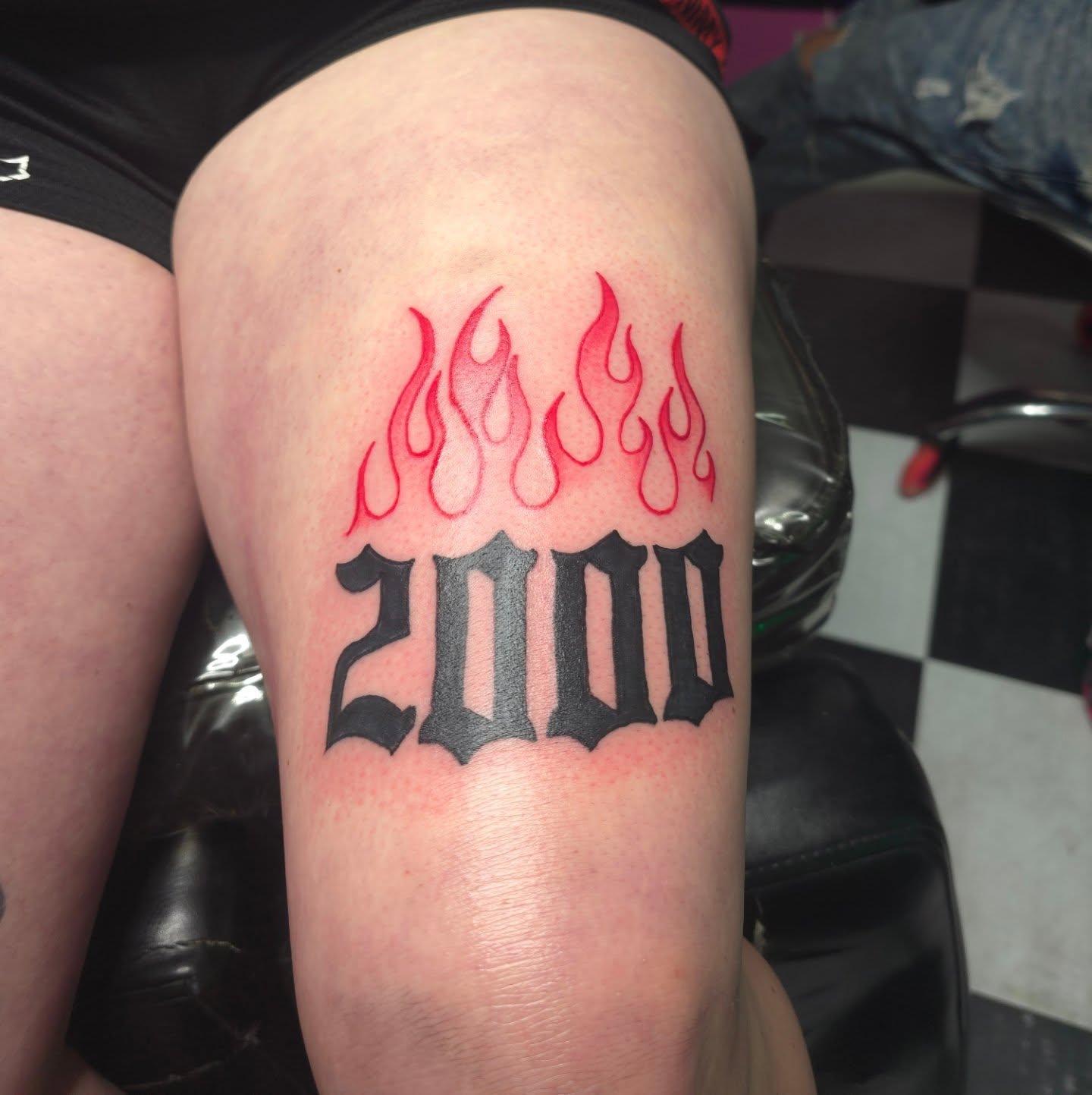 Flames and '2000' black and grey tattoo on the thigh by Kennedy Novella