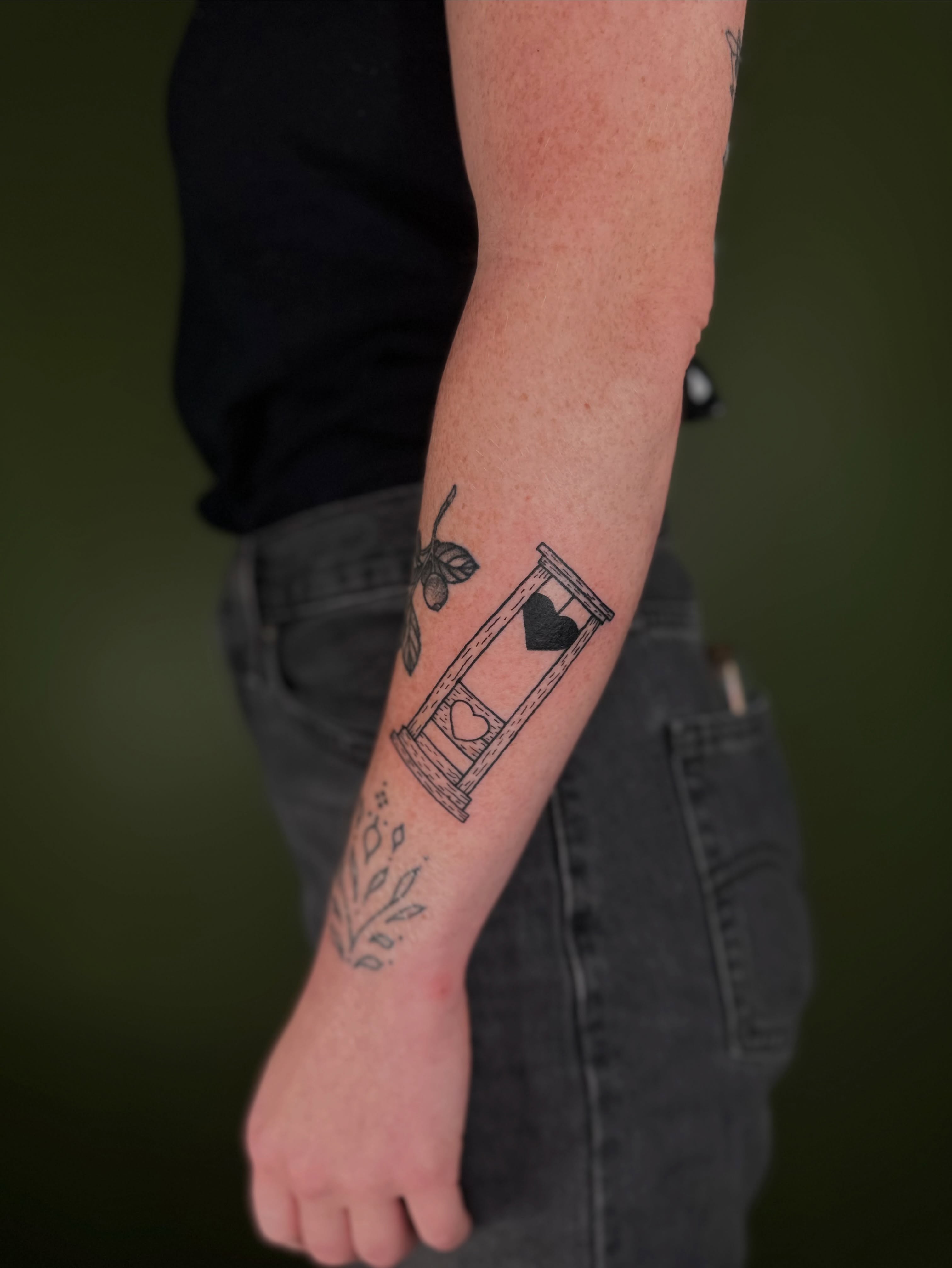 Hourglass with heart black and grey tattoo on the forearm by Nikolas Tapio Aatos Mäkelä
