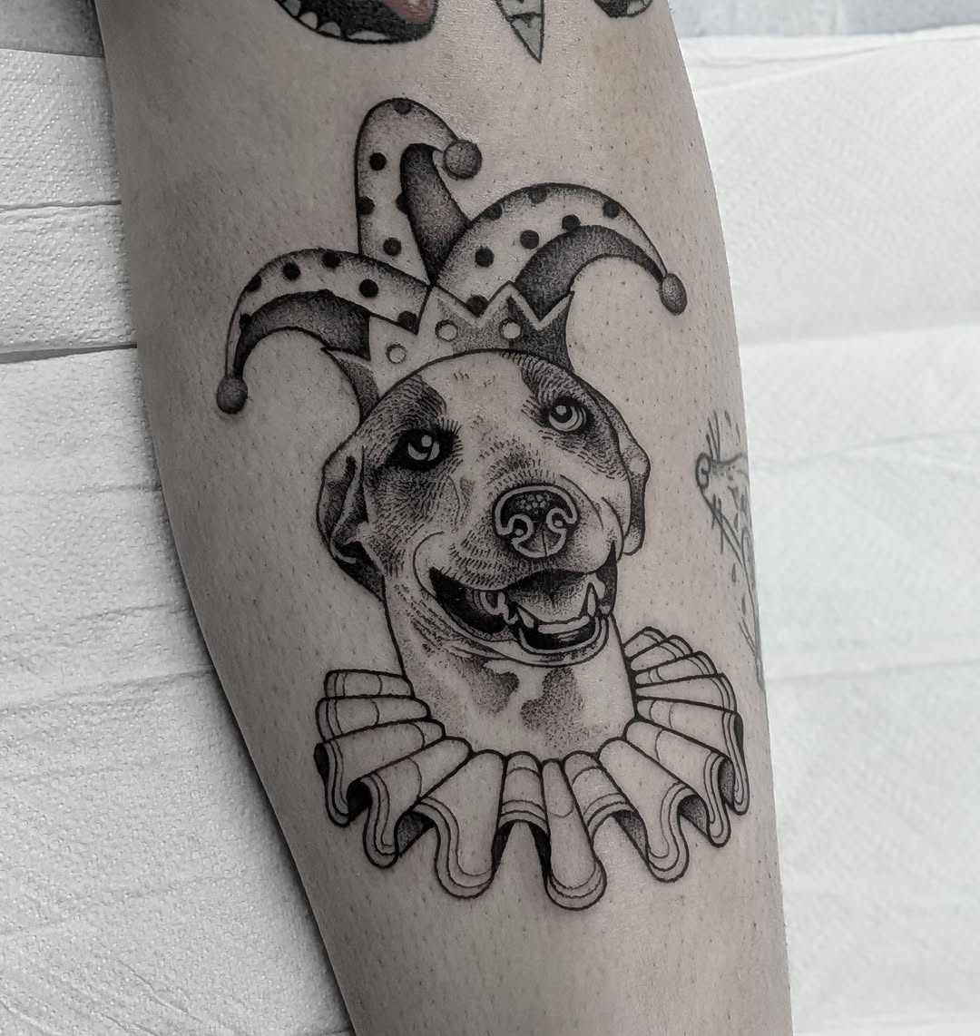 Dog in jester costume realism tattoo on the forearm by Joshua Harkey