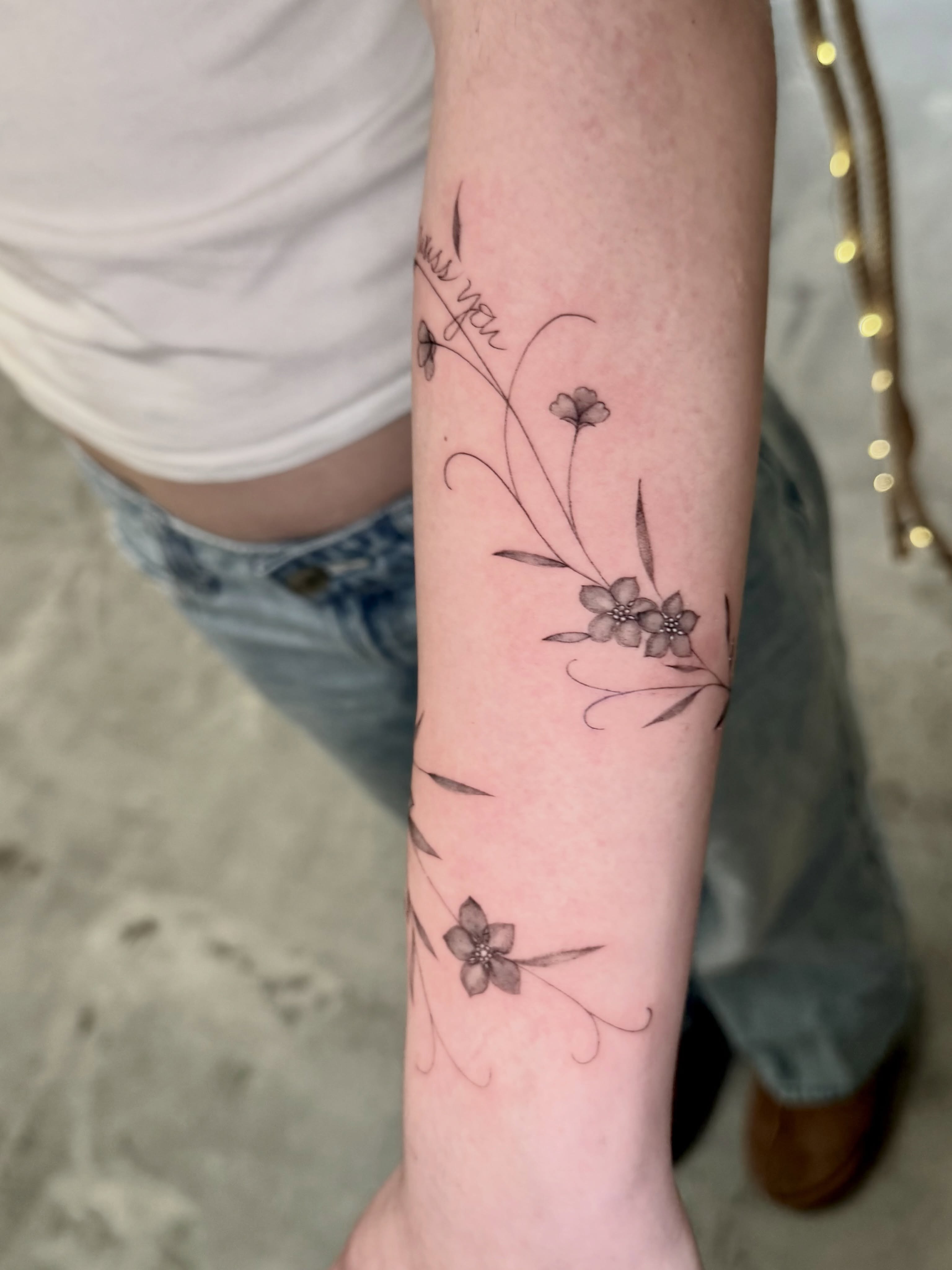 Flowers and script fine line tattoo on the forearm by Anna
