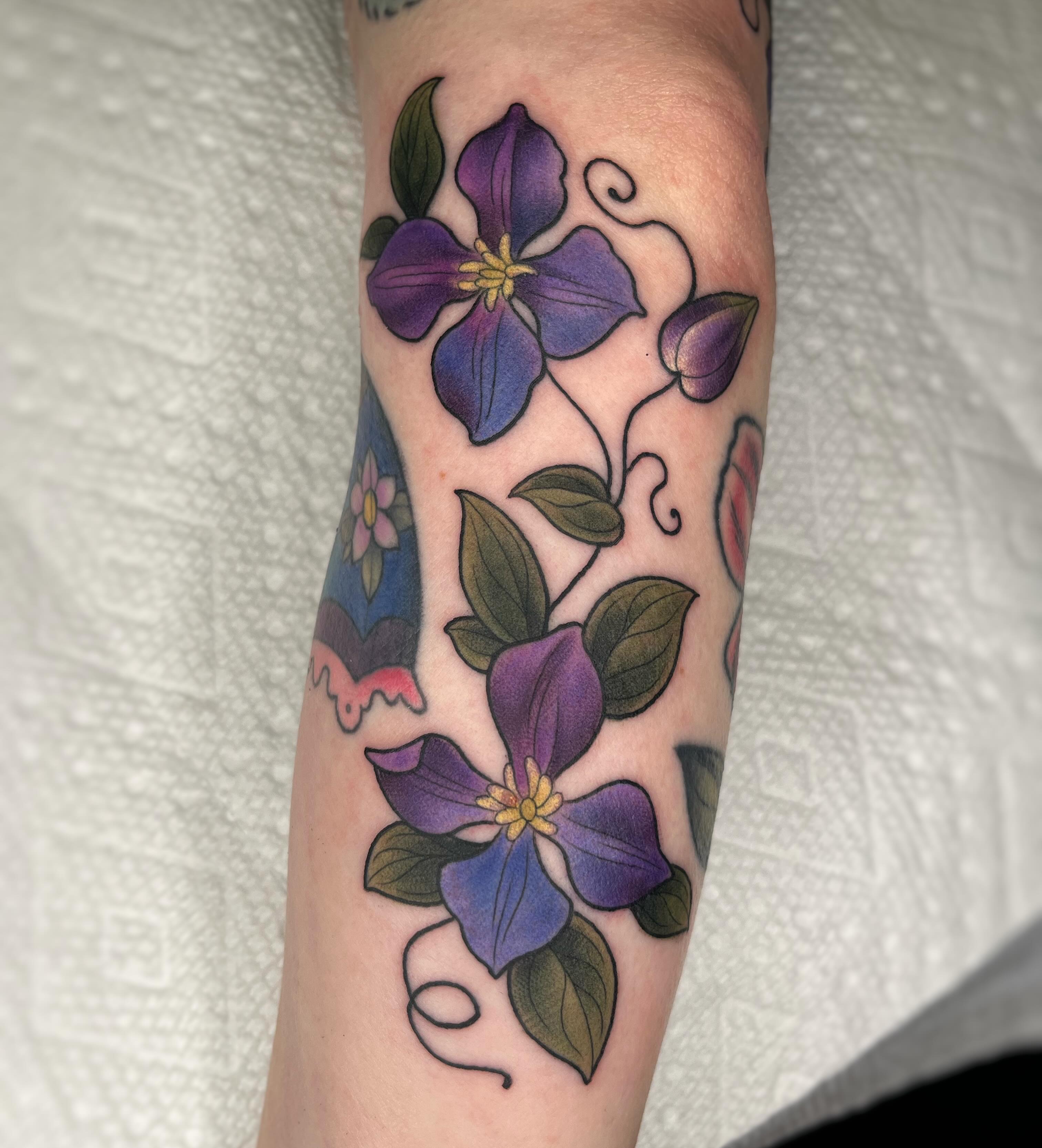Purple clematis flowers American traditional tattoo on the forearm by Emi Collison