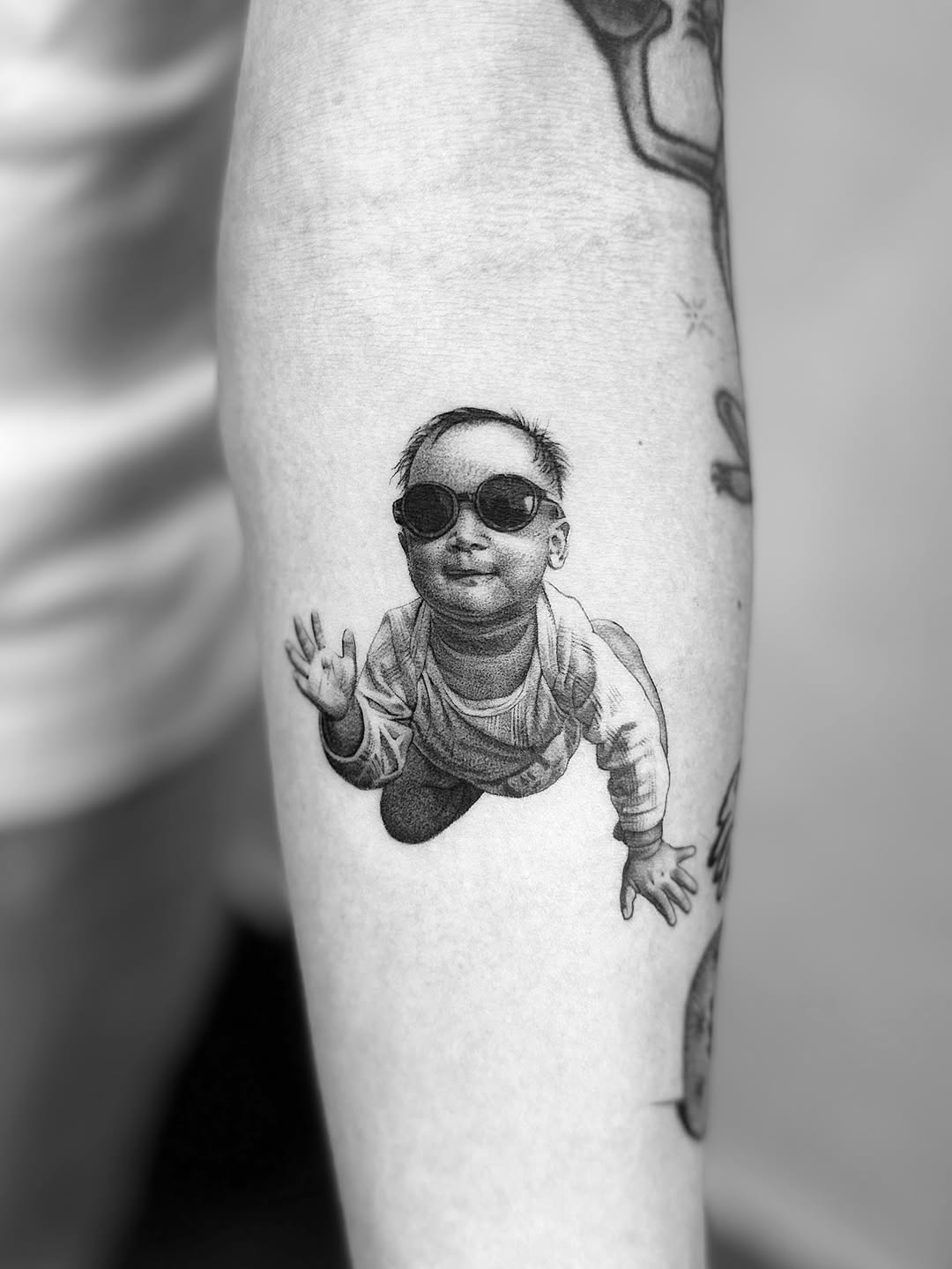 Baby wearing sunglasses realism tattoo on the forearm by Saru