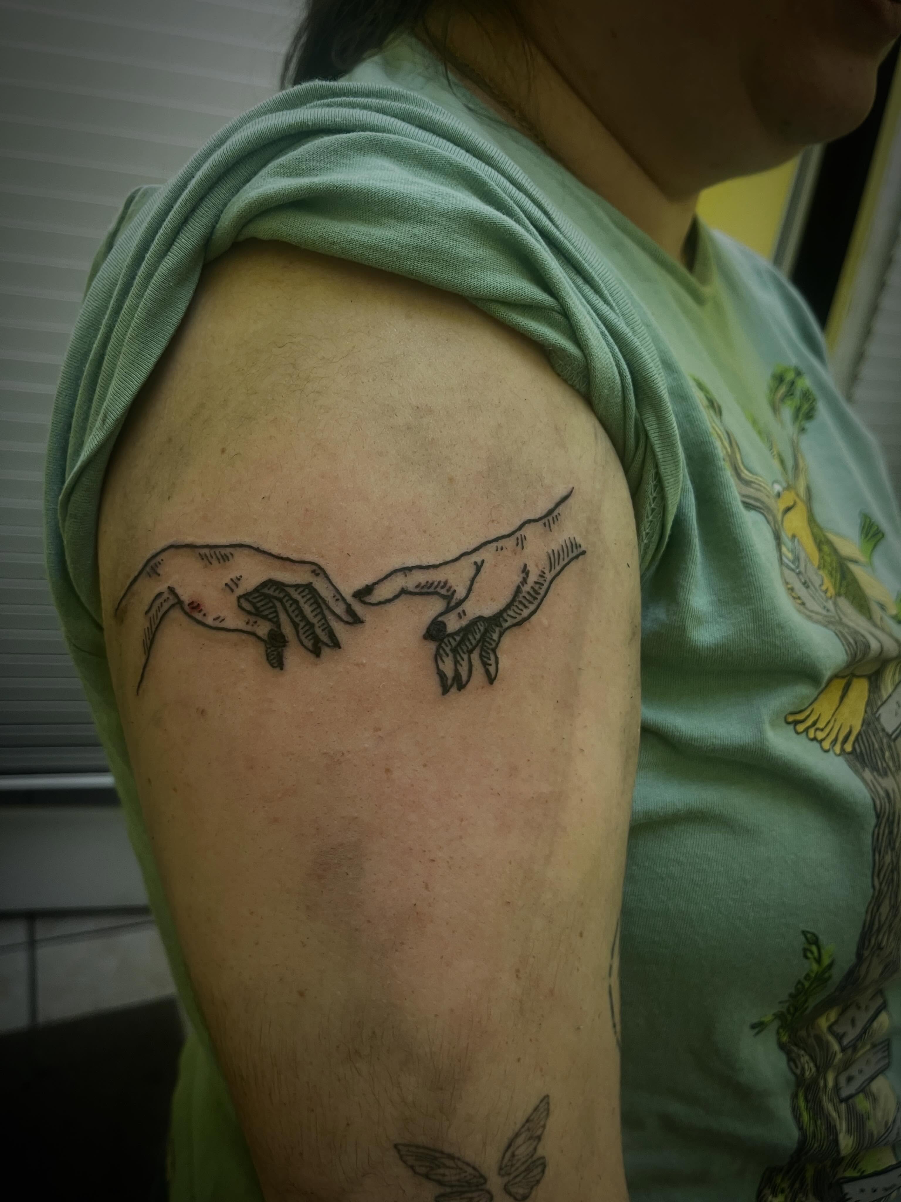Two hands touching black and grey tattoo on the shoulder by Rafaela Lopez
