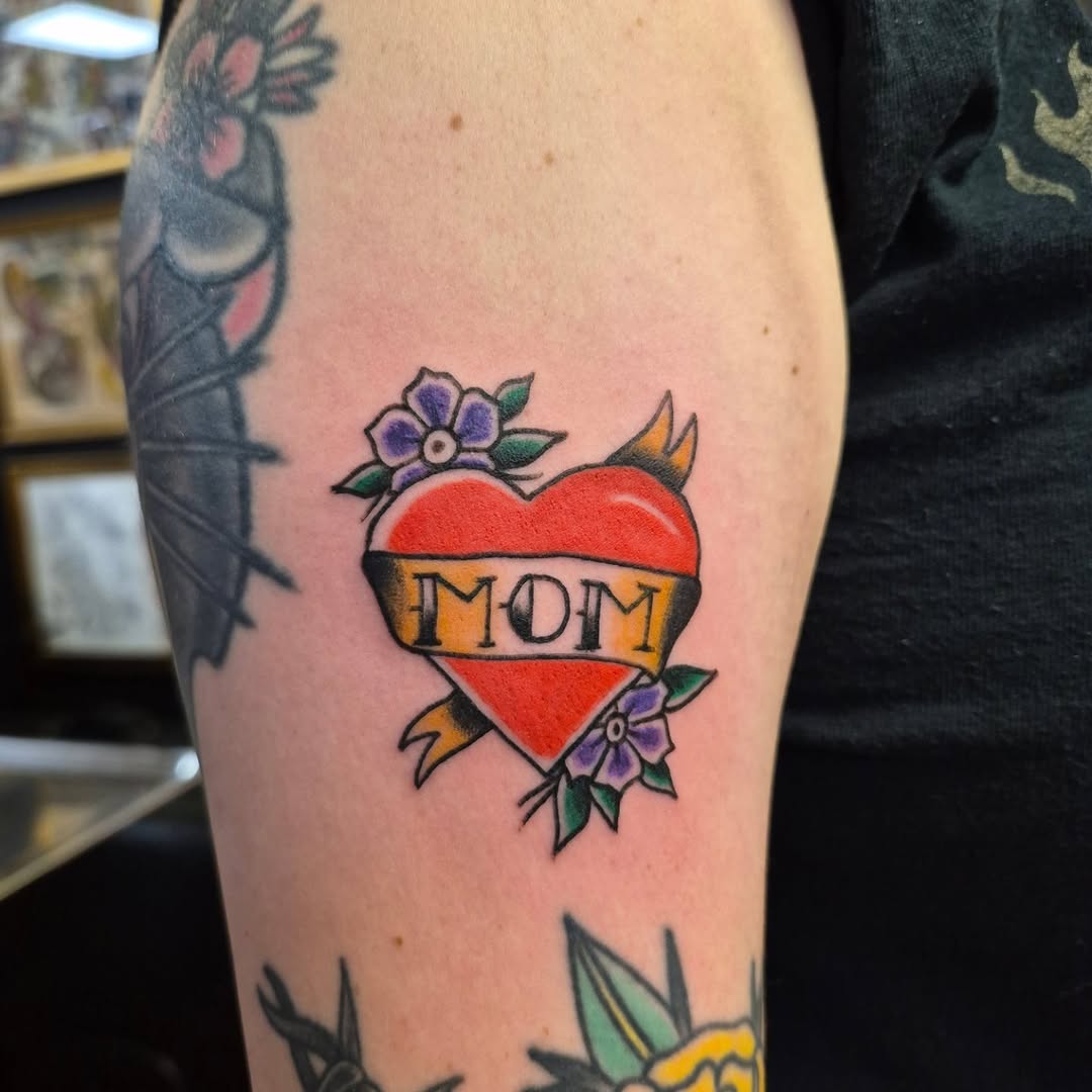 Heart with "MOM" banner American traditional tattoo on the forearm by Bobby H