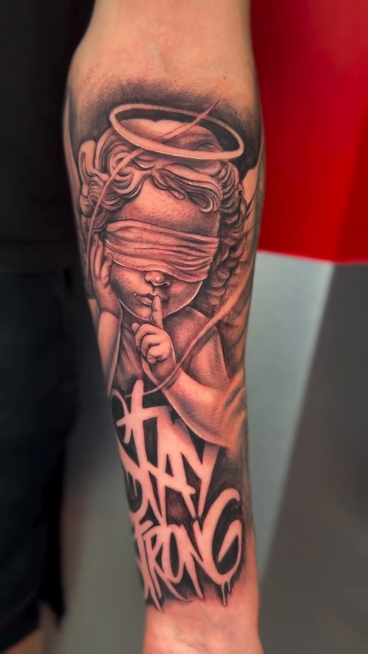 Blindfolded cherub with "Stay Strong" lettering realism tattoo on the forearm by guinn_art_tatts, Le Moule