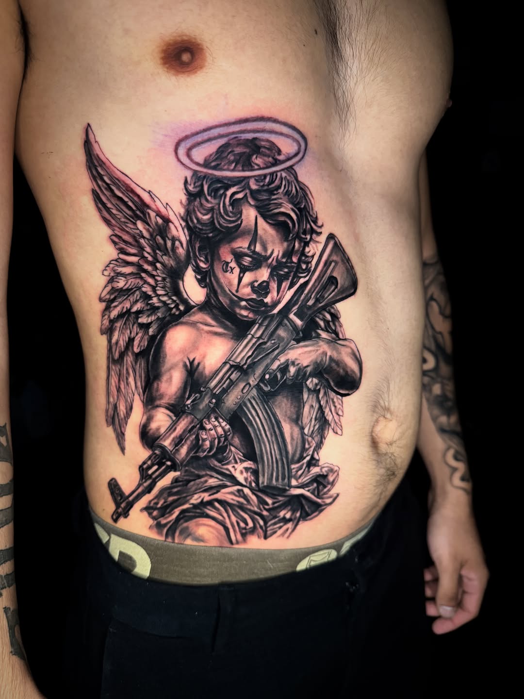 Cherub with gun and clown makeup realism tattoo on the ribs by Dallas Tattoo Artist📍