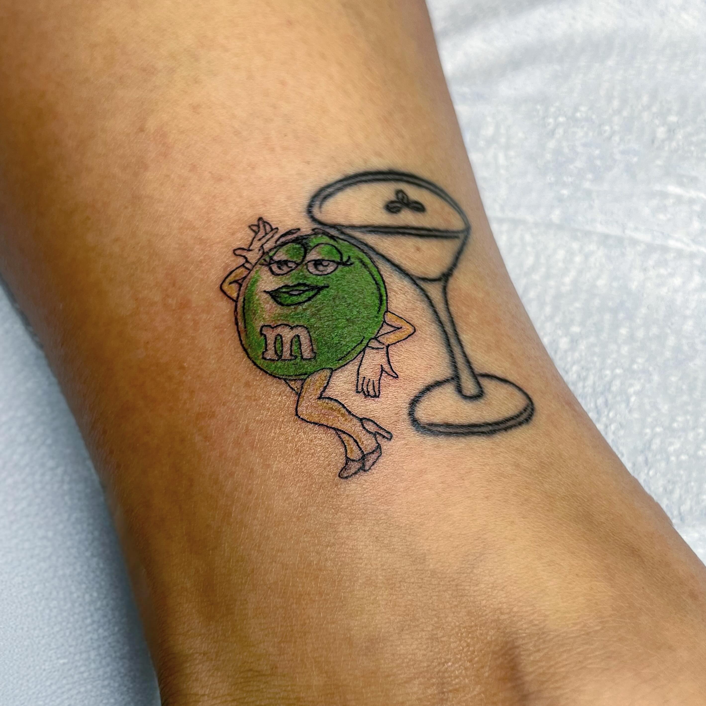Green M&M character illustrative tattoo on the forearm by Sara Atamian, San Diego, California