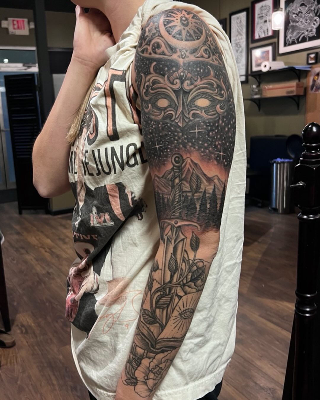 Mask and landscape neo-traditional tattoo on the upper arm by Jesse Gray