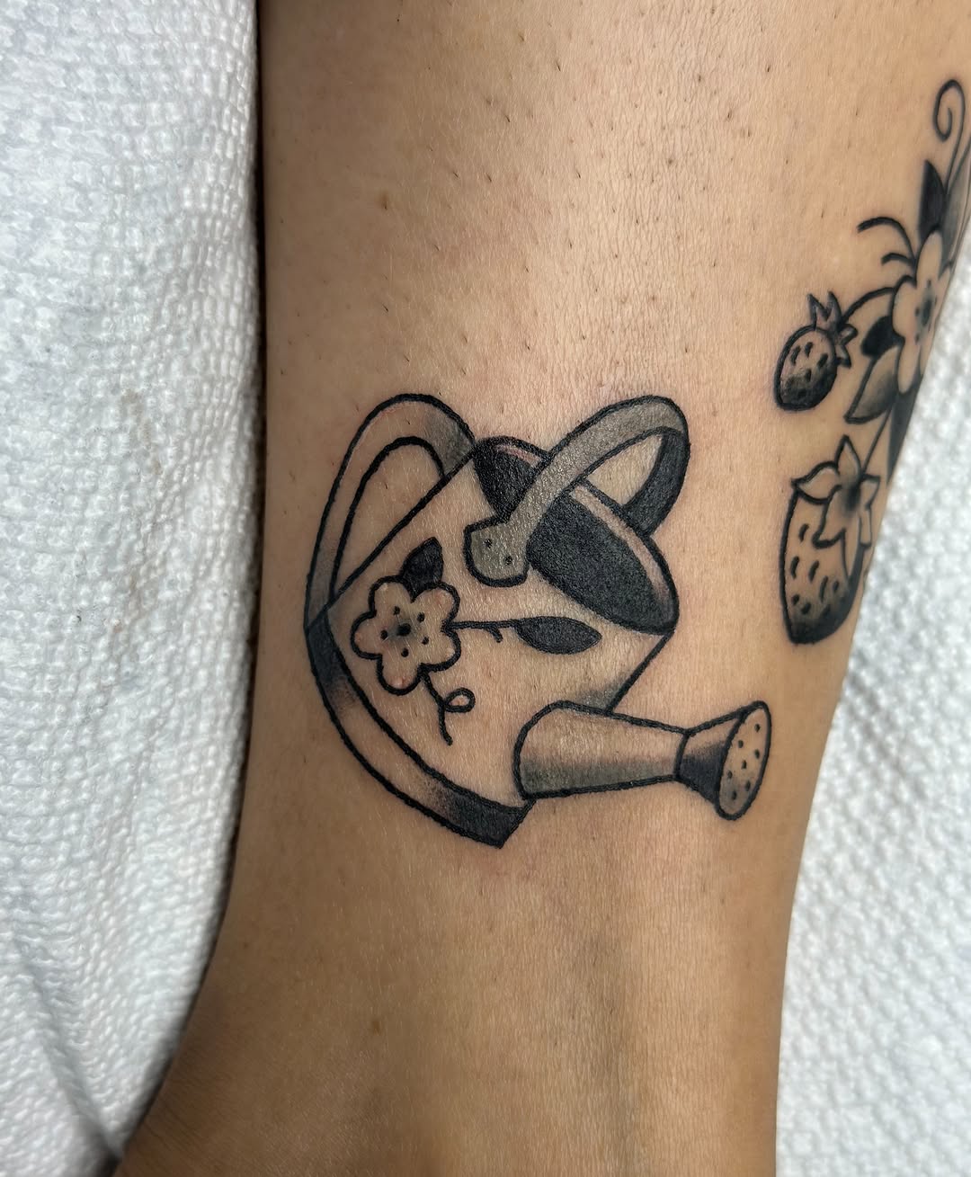 Watering can with flower and strawberries minimalist tattoo on the forearm by Lucy Jay
