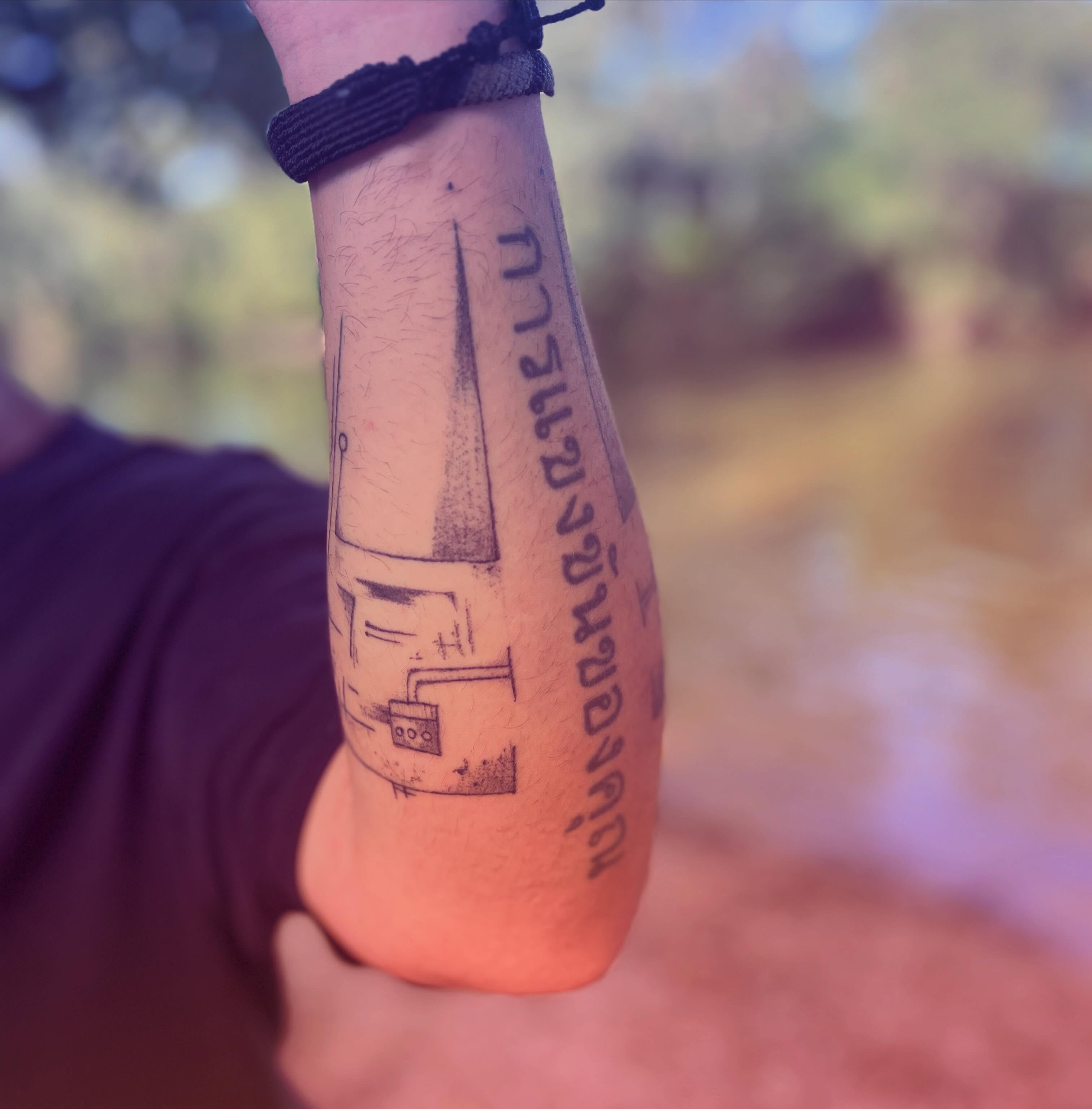 Building and Thai script black and grey tattoo on the forearm by La Main Bleue