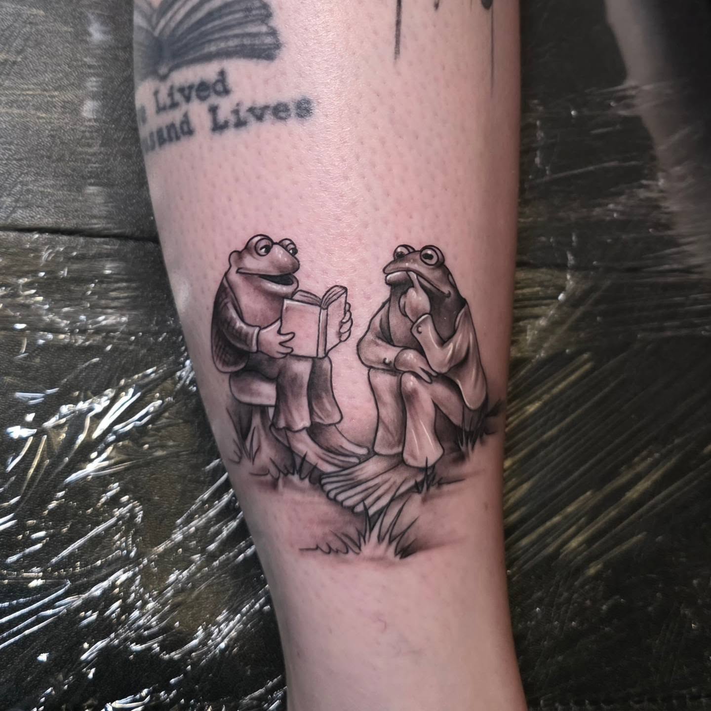Frogs reading realism tattoo on the forearm by Kosta