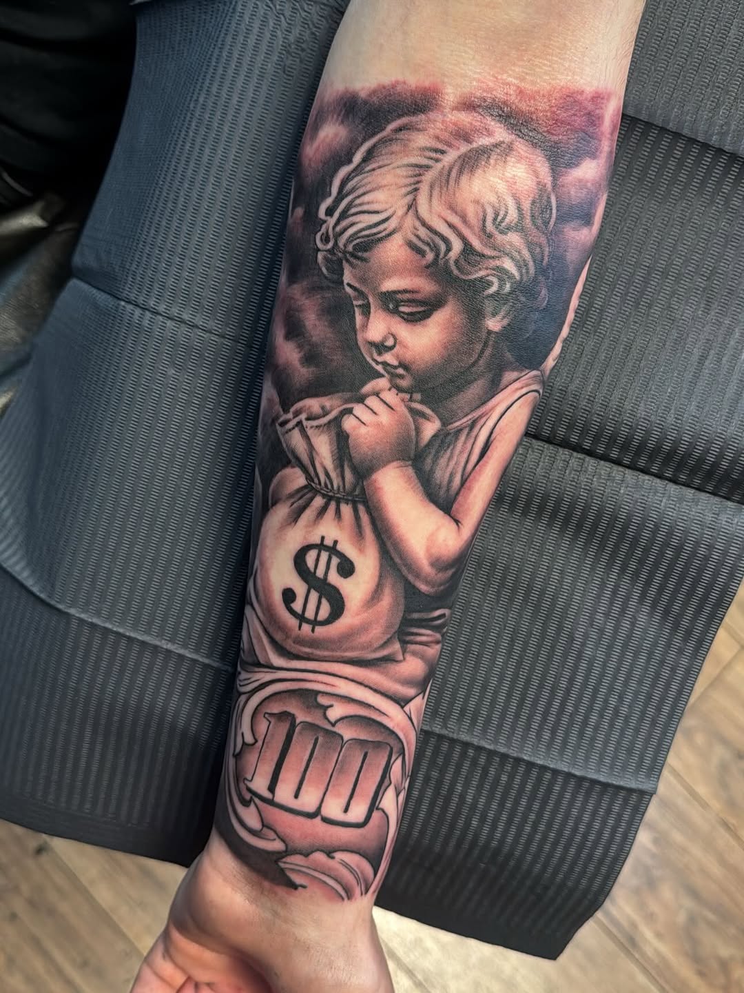 Cherub holding money bag with 100 realism tattoo on the forearm by Cedric Fleming