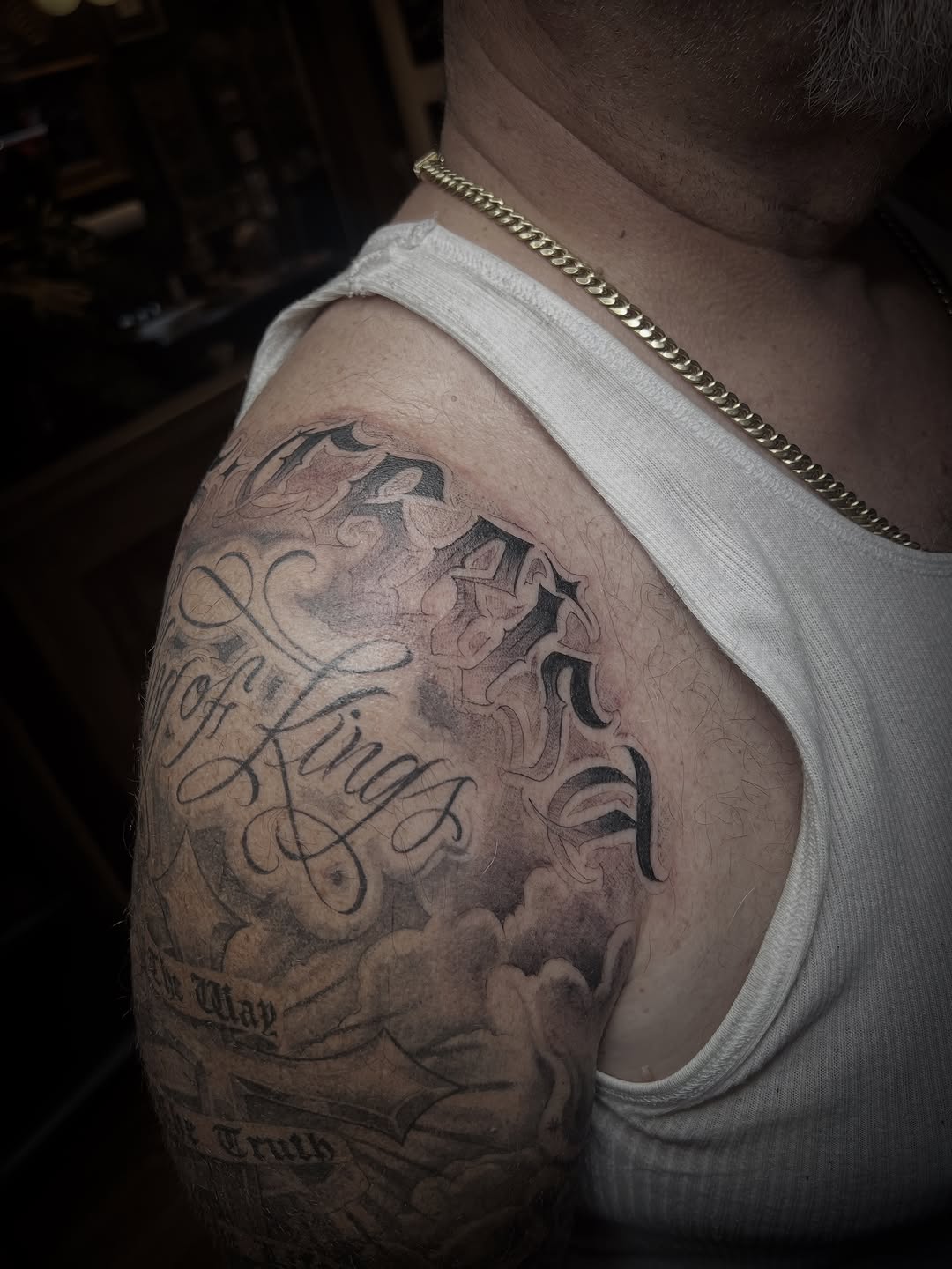 Script lettering with clouds Chicano tattoo on the shoulder by Oso