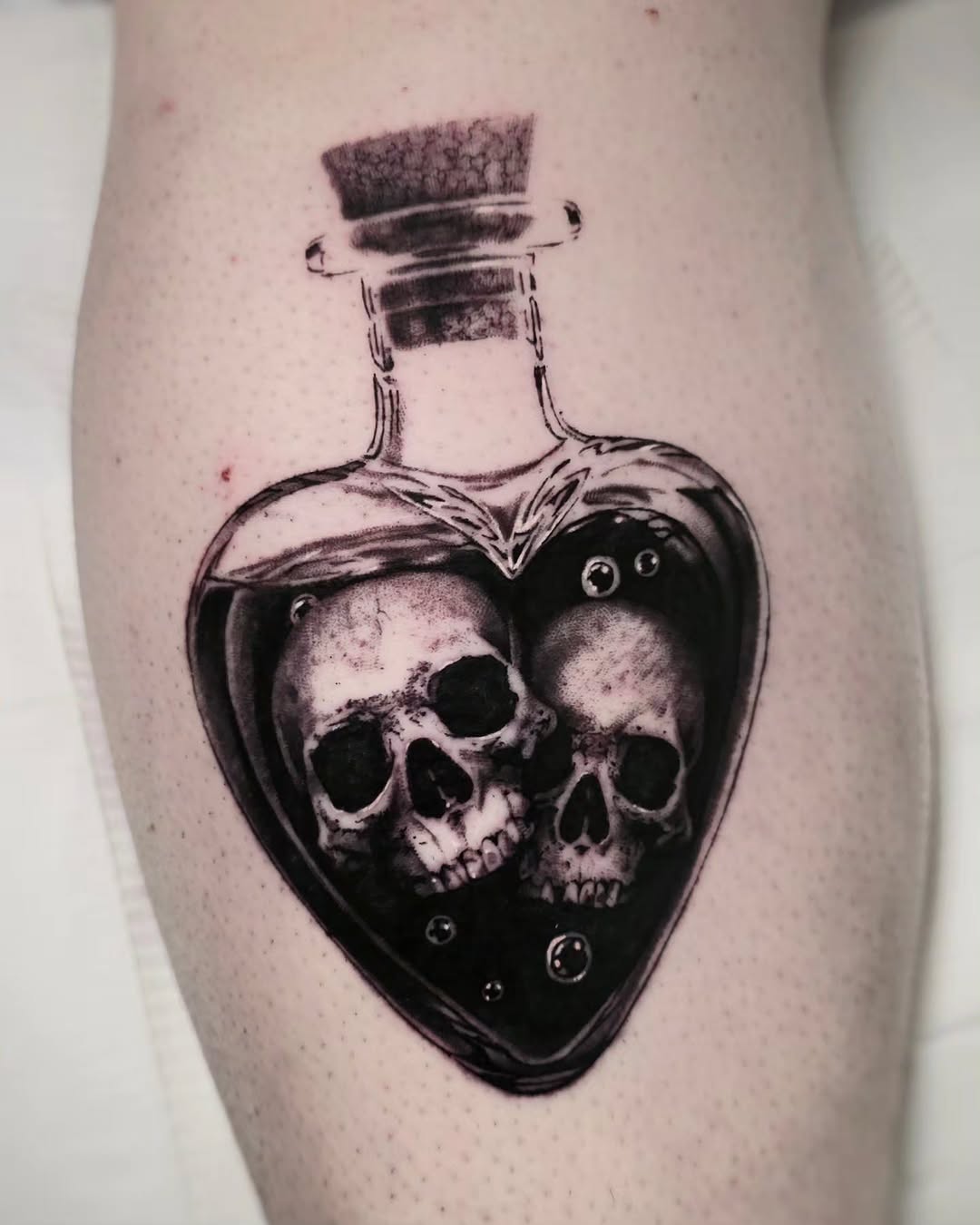 Two skulls in a heart-shaped bottle realism tattoo on the thigh by ʙʀɪᴀɴɴᴀ 𝒁𝑶𝑴𝑩𝑹𝑰𝑬 ᴛᴀᴛᴛᴏᴏꜱ