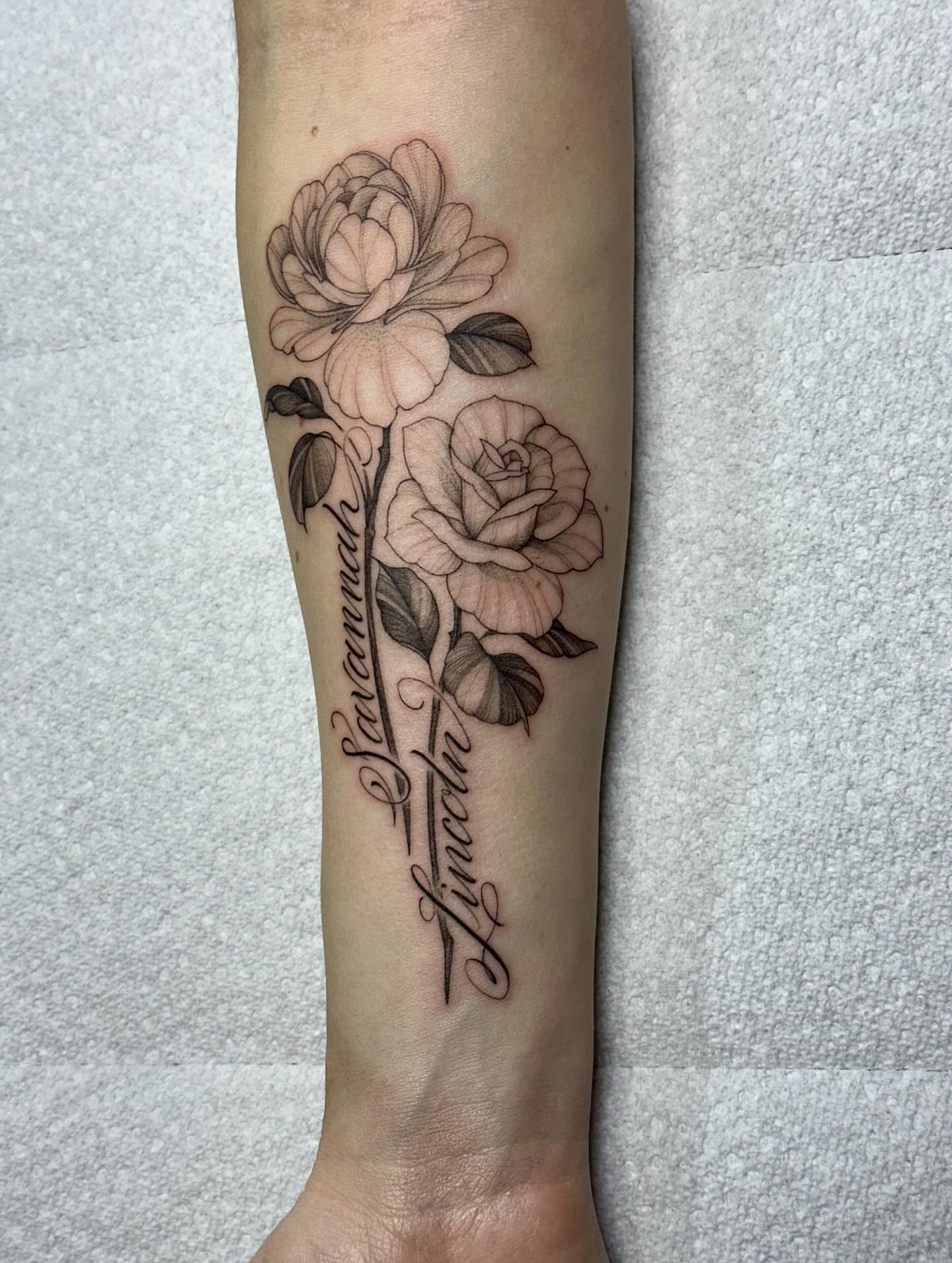 Flowers and names black and grey tattoo on the forearm by Izabelle Inks