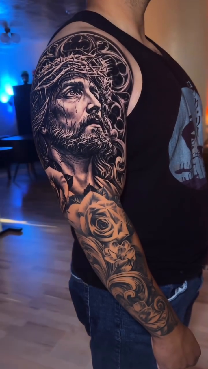 Jesus Christ portrait with roses Chicano tattoo on the upper arm by 𝐉𝐨𝐧 𝐒𝐚𝐧𝐭𝐨𝐬