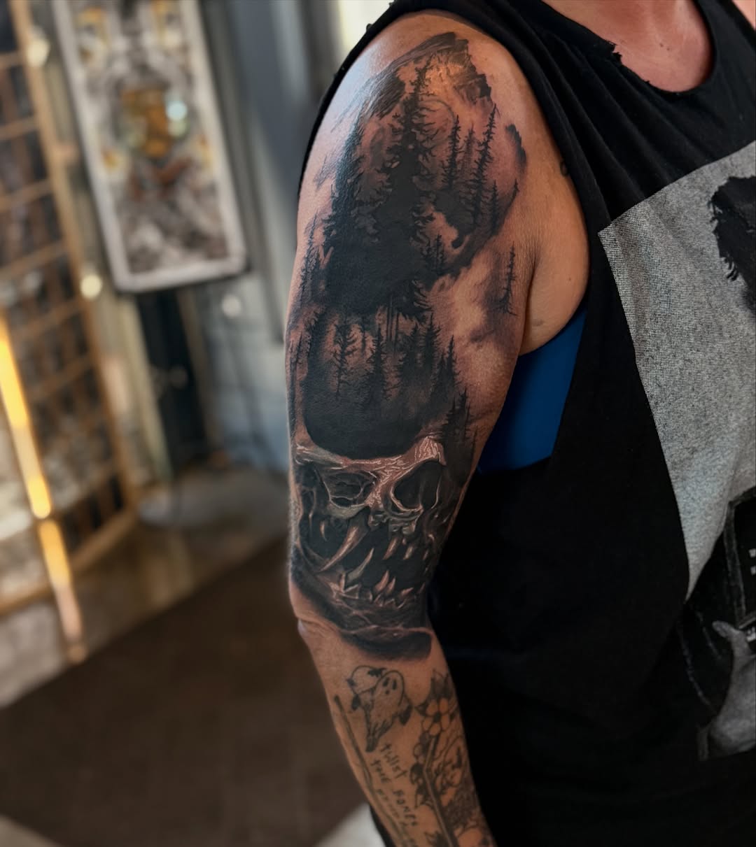Skull with forest and moon dark art tattoo on the upper arm by Miles Haysmith