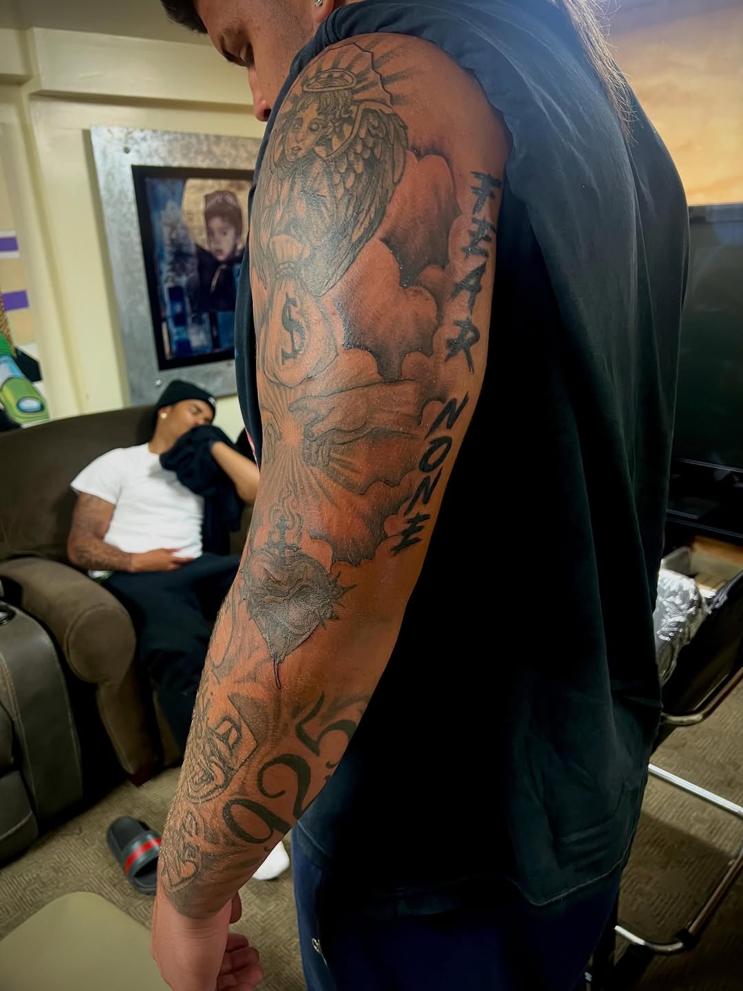 Angel, money bags, clouds, lettering Chicano tattoo on the upper arm by Jelani