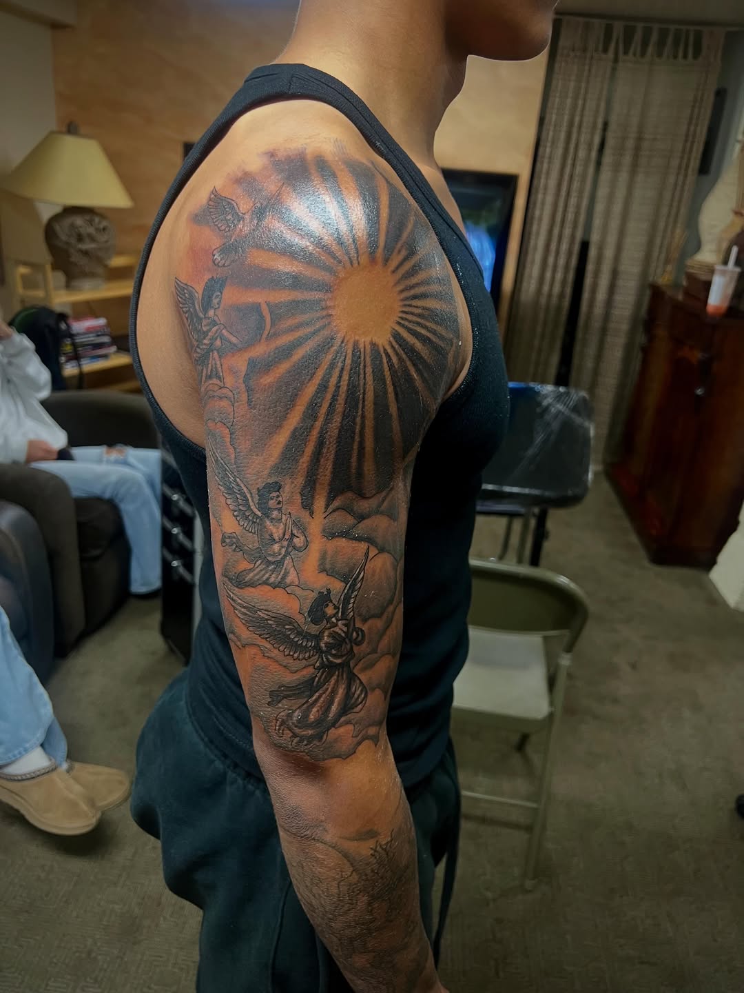 Sun with rays and angels realism tattoo on the upper arm by Jelani