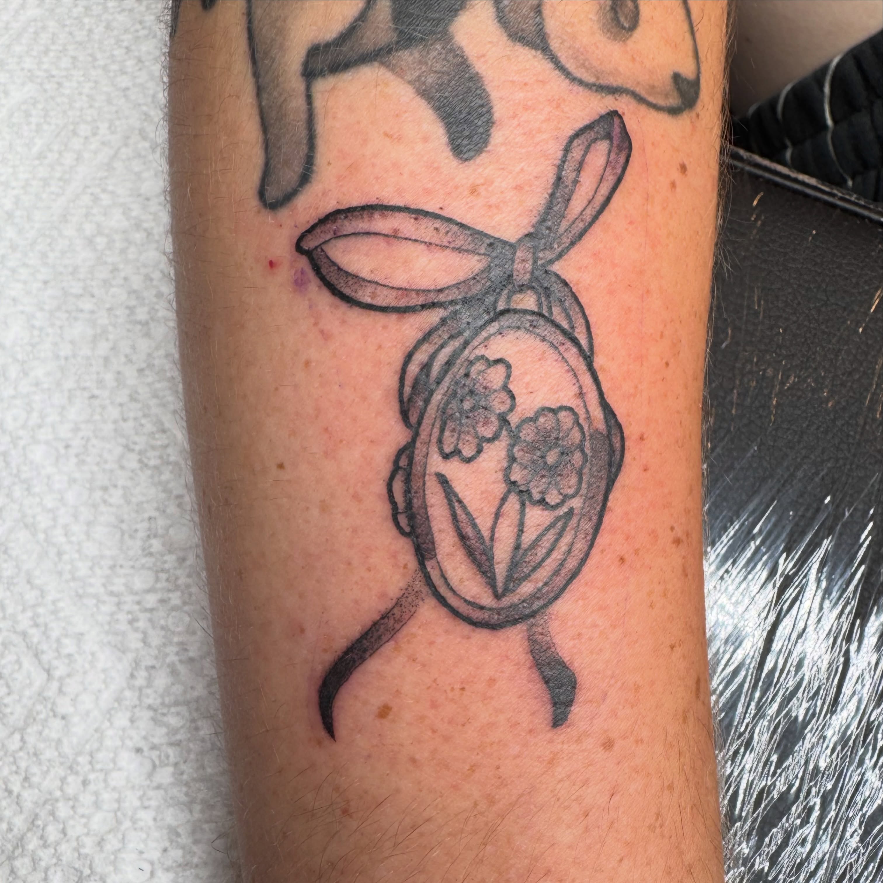 Locket with flowers black and grey tattoo on the forearm by tori 🫶🏻
