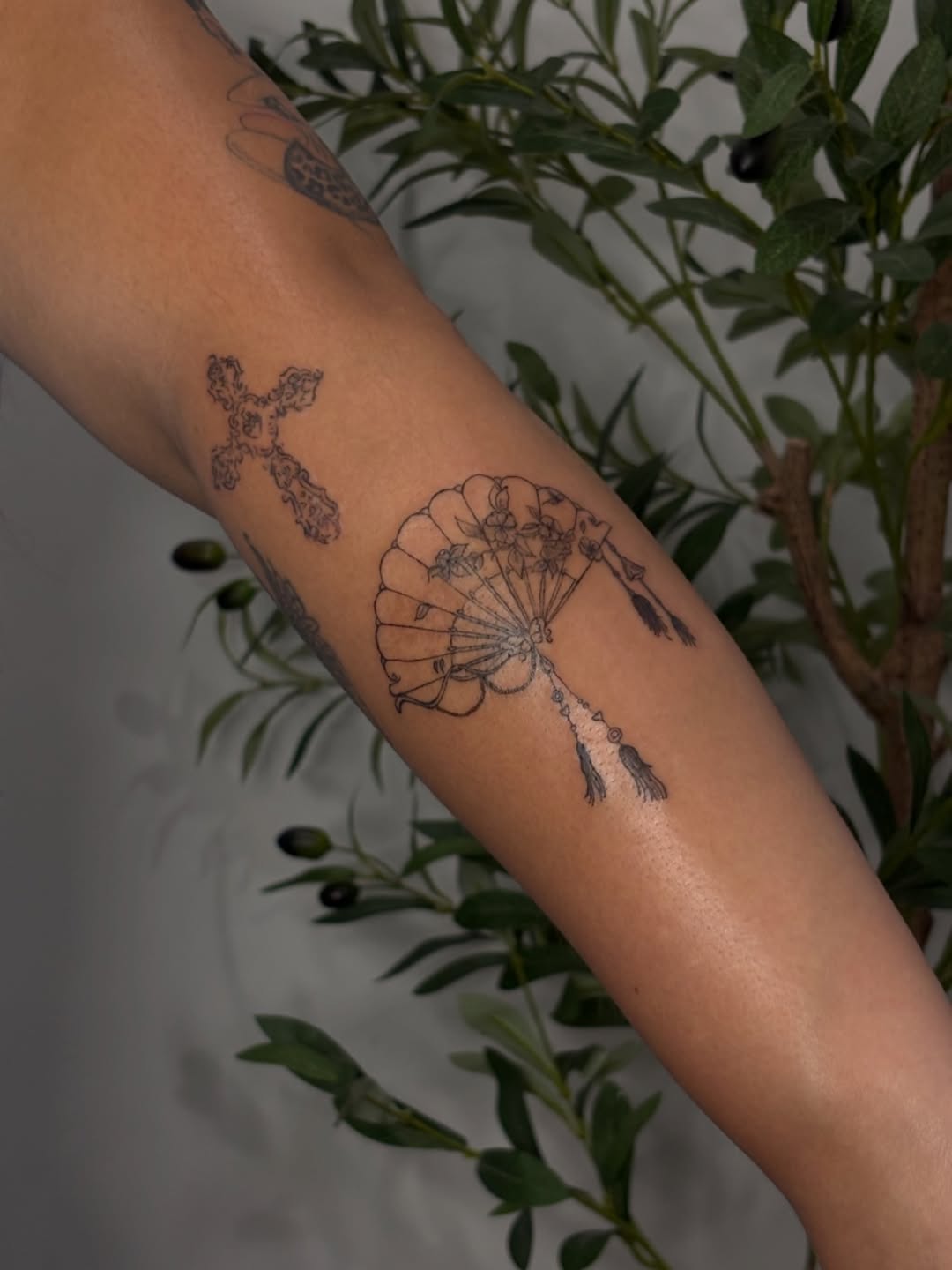 Ornate fan and cross minimalist tattoo on the forearm by Valentina