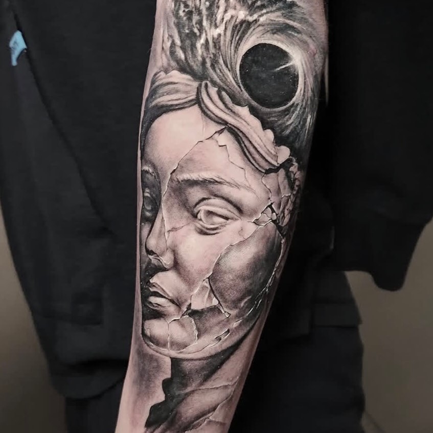 Cracked statue head with black hole surrealism tattoo on the forearm by Elidia Veliz