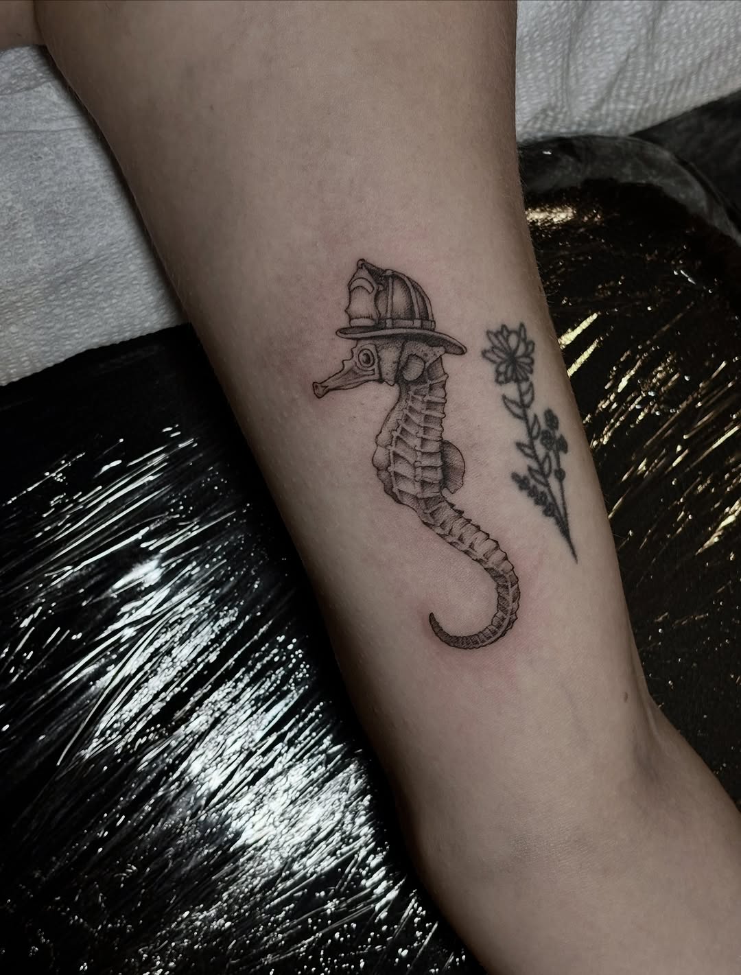 Seahorse wearing a firefighter helmet fine line tattoo on the forearm by Emma Cotter