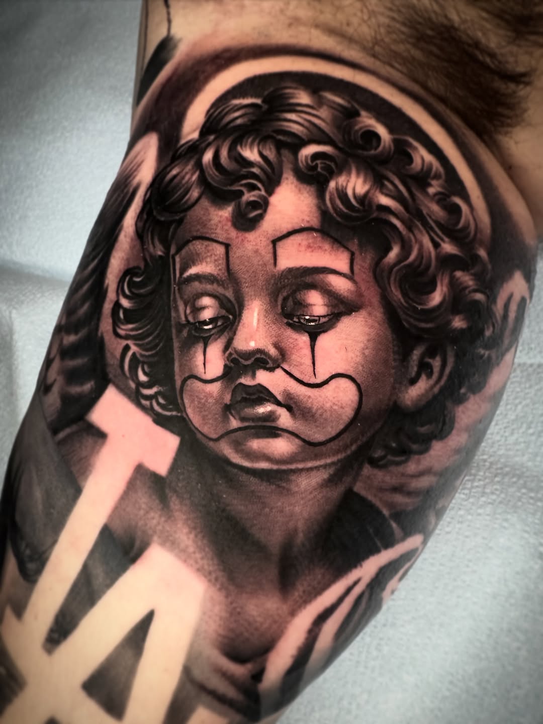 Sad clown cherub with LA logo Chicano tattoo on the forearm by David Torres