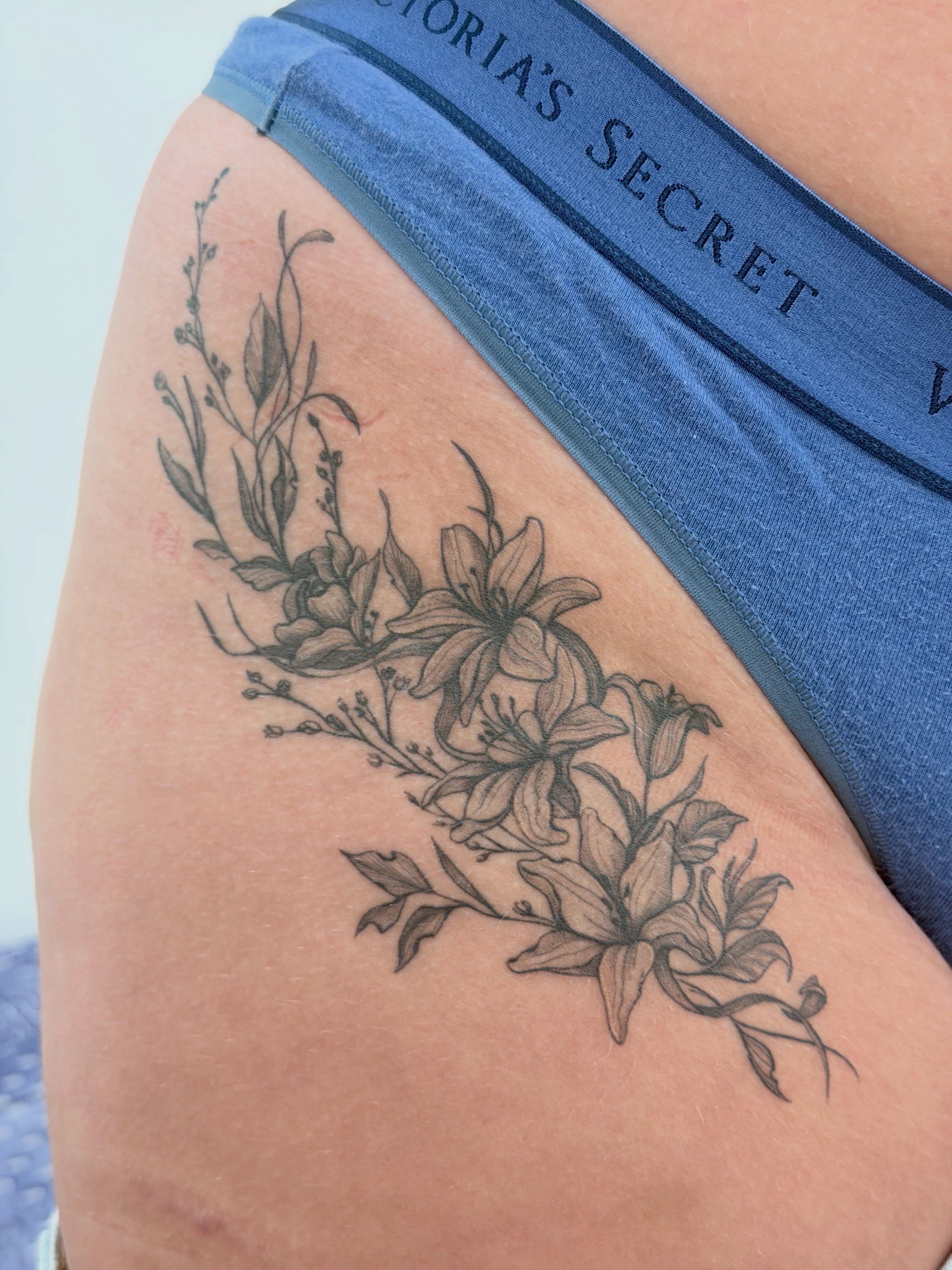 Lilies and leaves black and grey tattoo on the thigh by Ink About Me - Amanda’s Fine Lines
