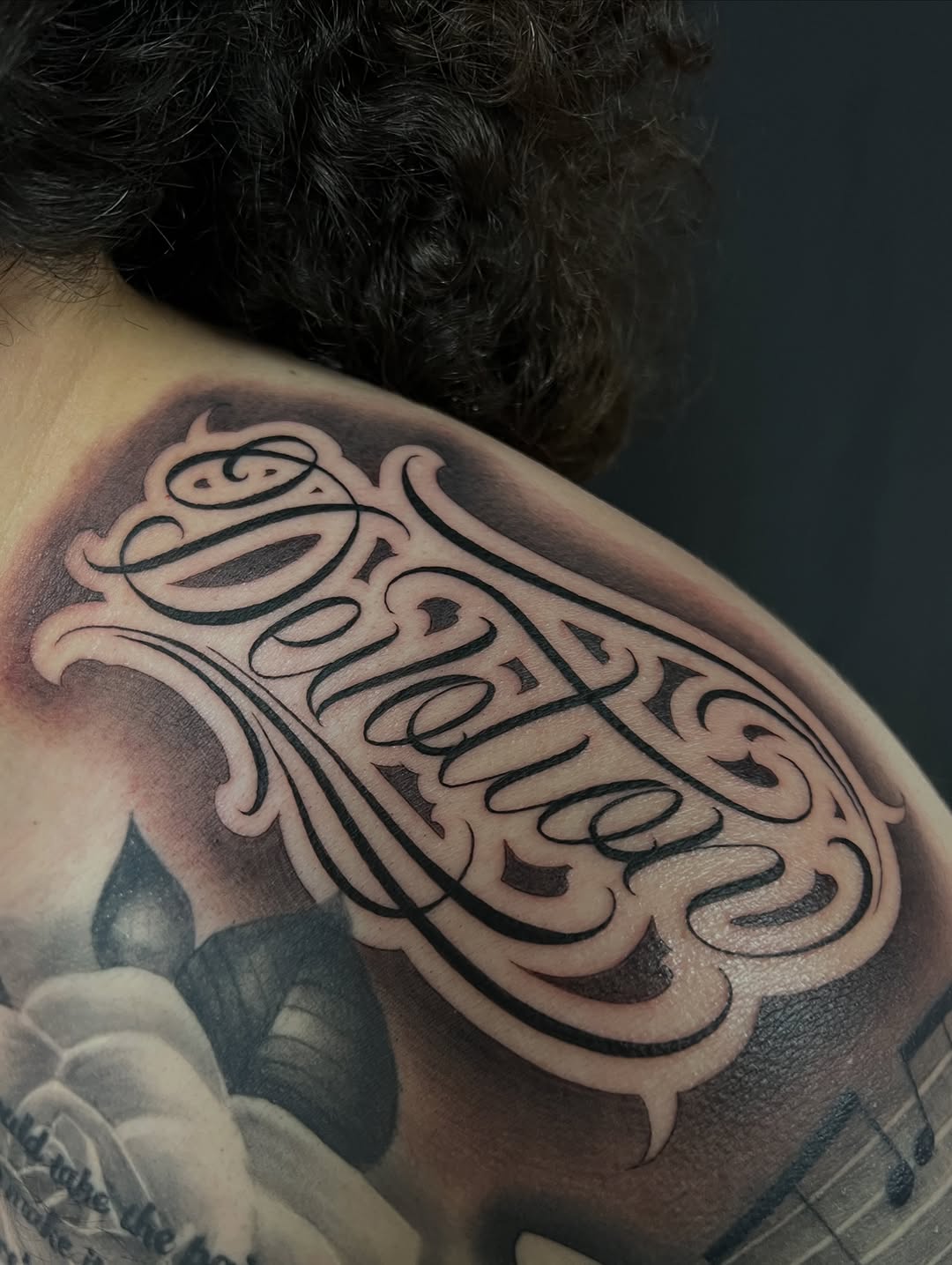 Ornamental script lettering Chicano tattoo on the shoulder by Thomas Conard