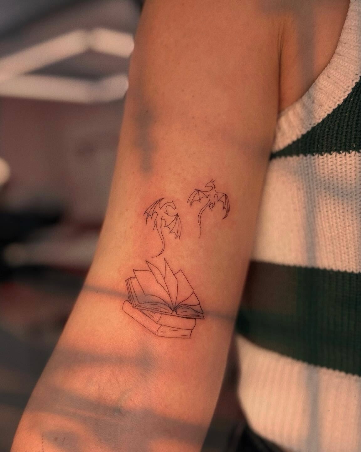 Book and dragons minimalist tattoo on the forearm by Ellie