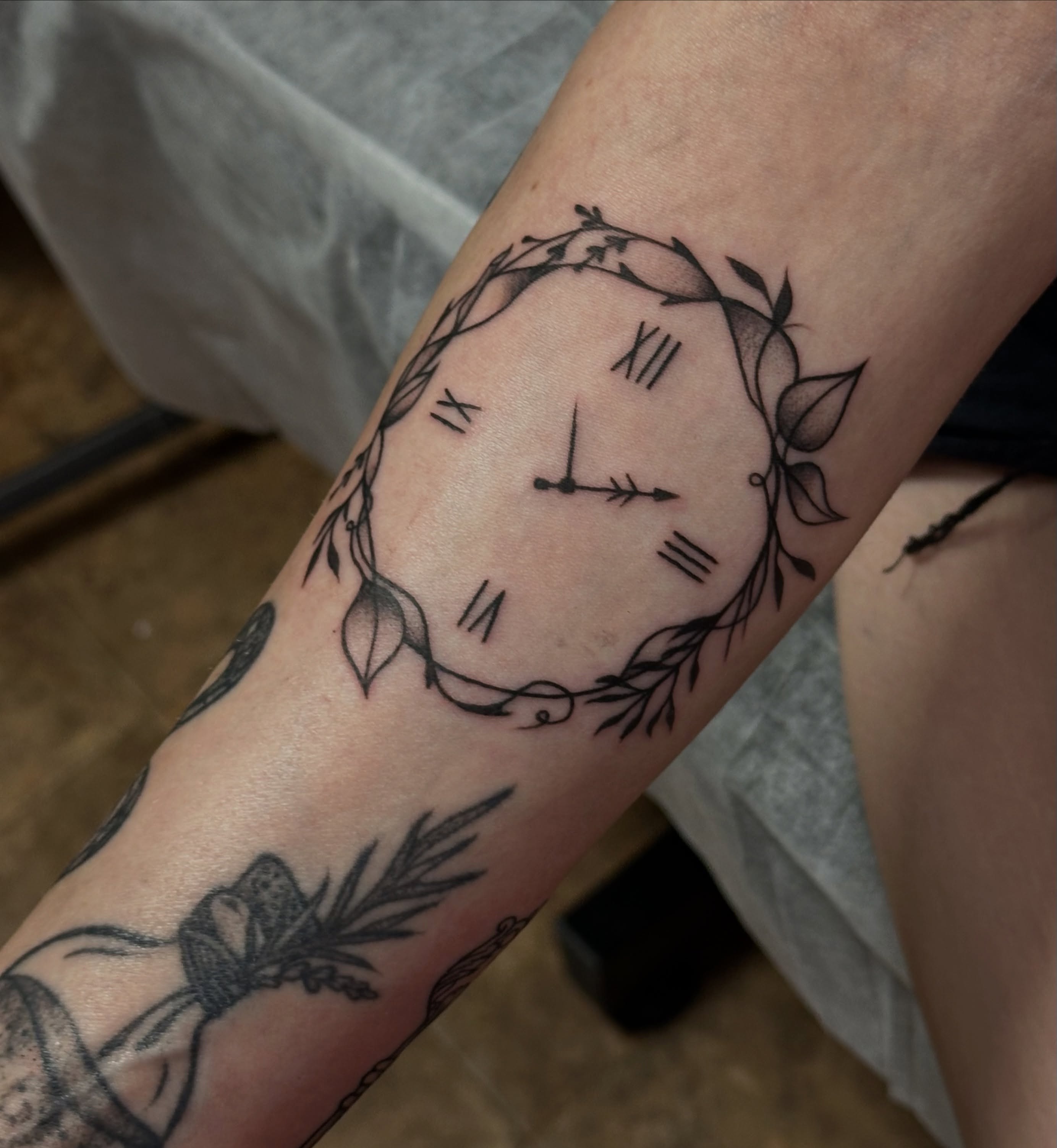 Clock with leaves black and grey tattoo on the forearm by Jocelyn