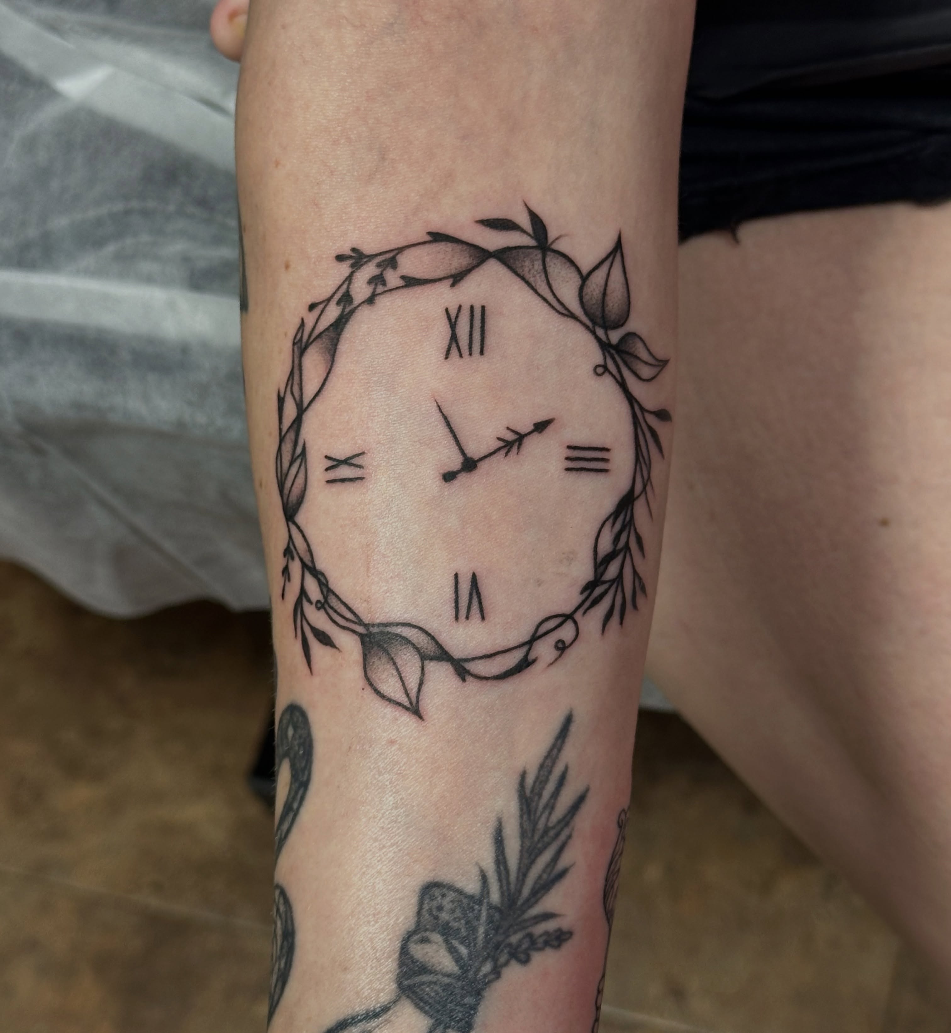 Clock with floral wreath black and grey tattoo on the forearm by Jocelyn