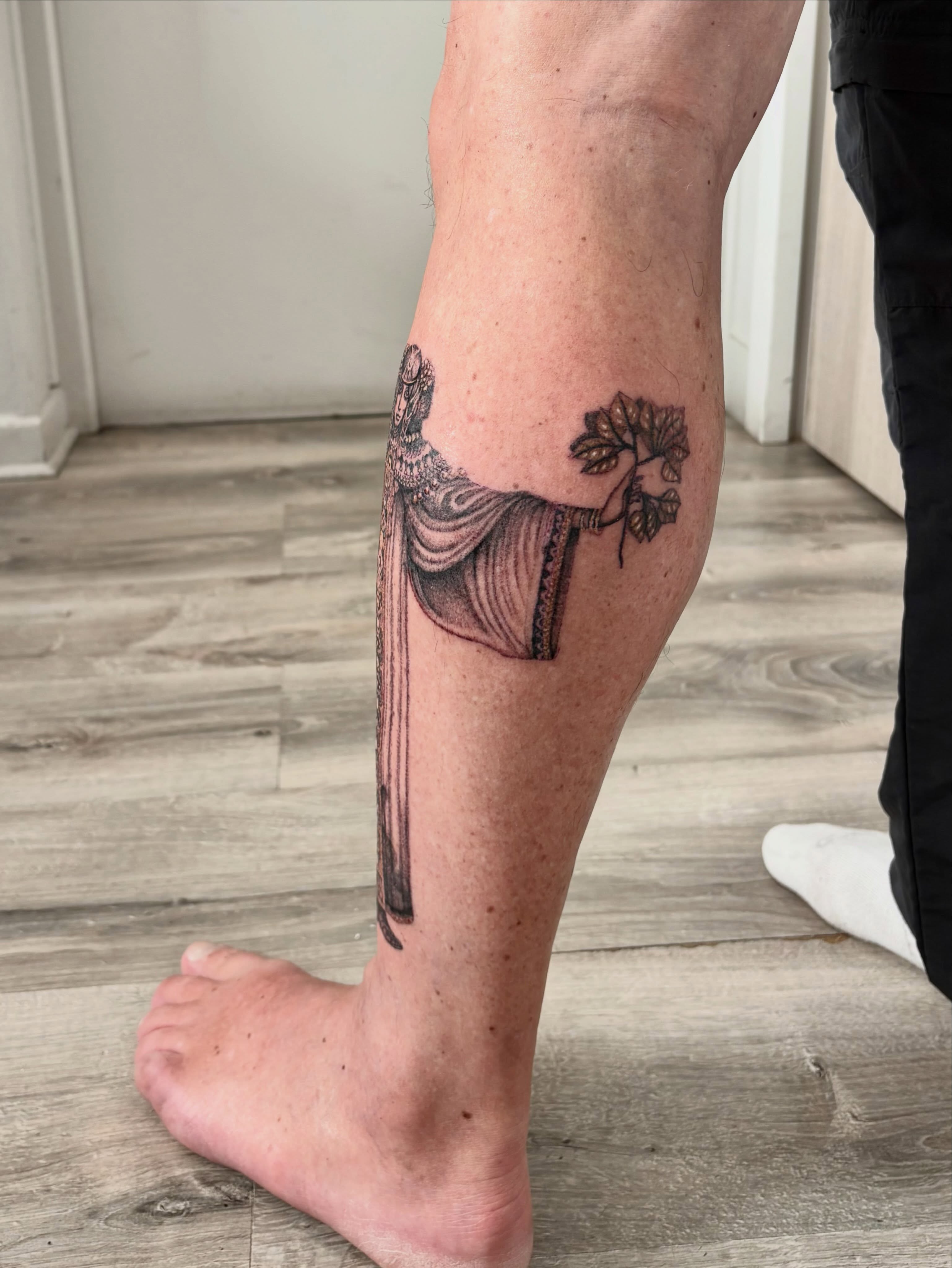 Draped figure with tree black and grey tattoo on the calf by Deborah