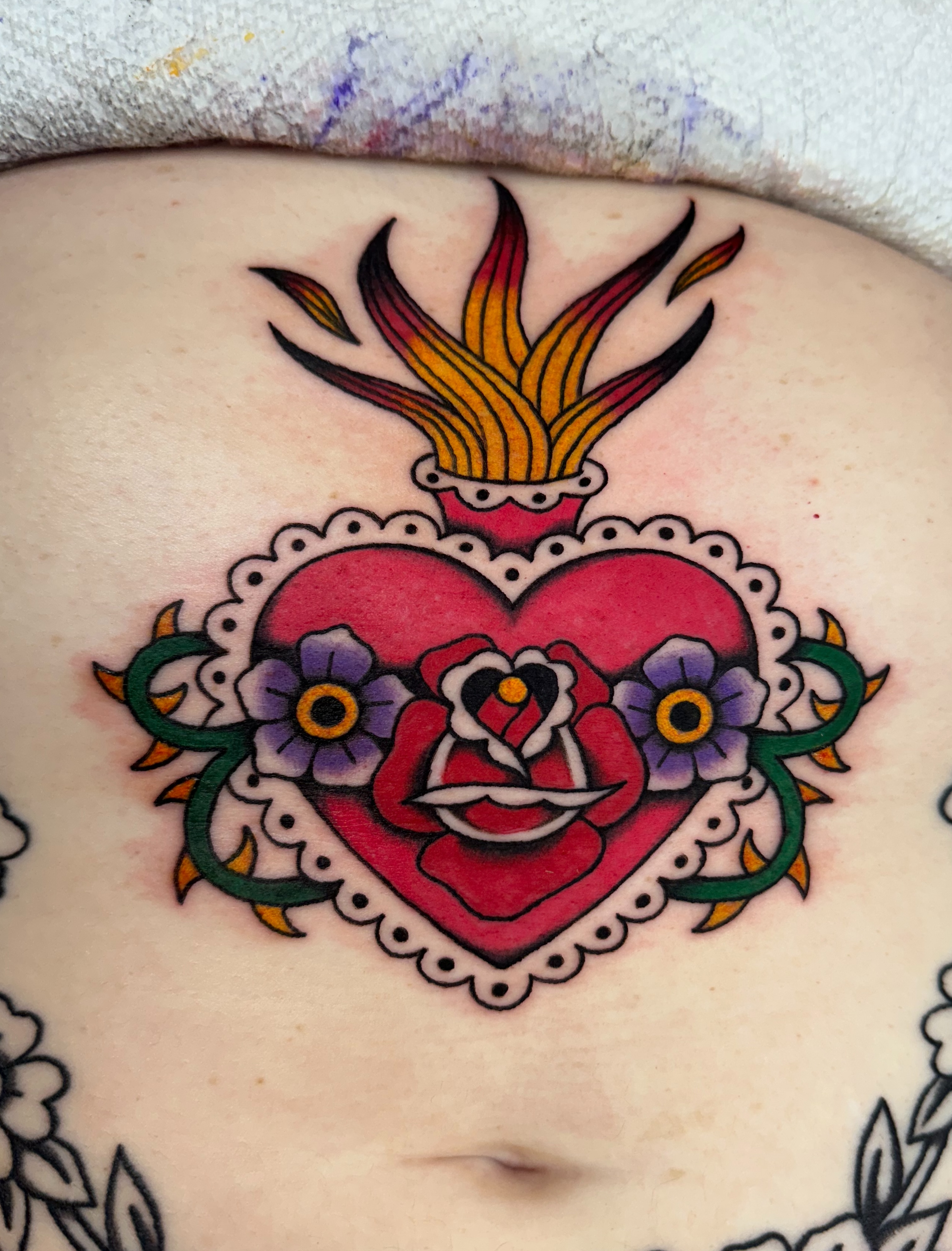 Heart with flames and flowers American traditional tattoo on the chest by Emily Masa