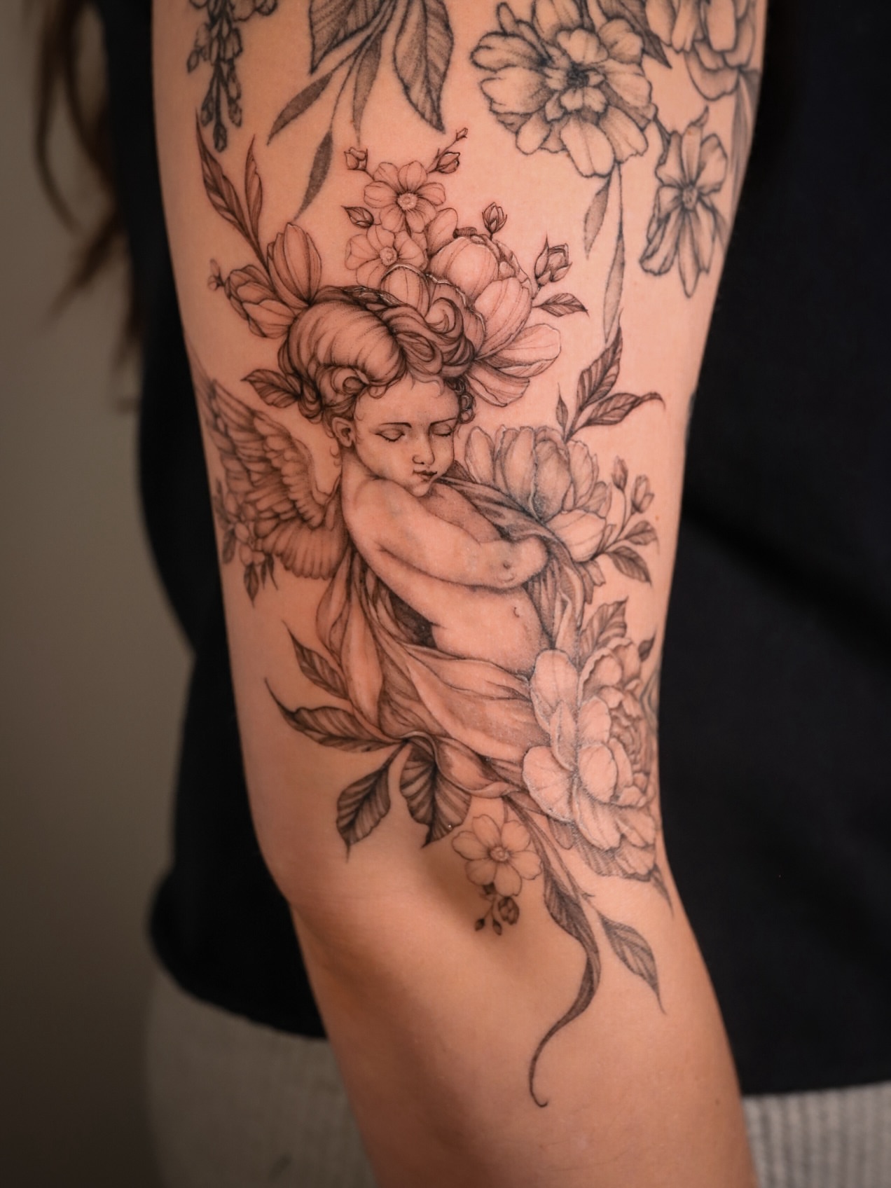 Cherub with flowers black and grey tattoo on the forearm by Caroline + Utah Tattoo Artist