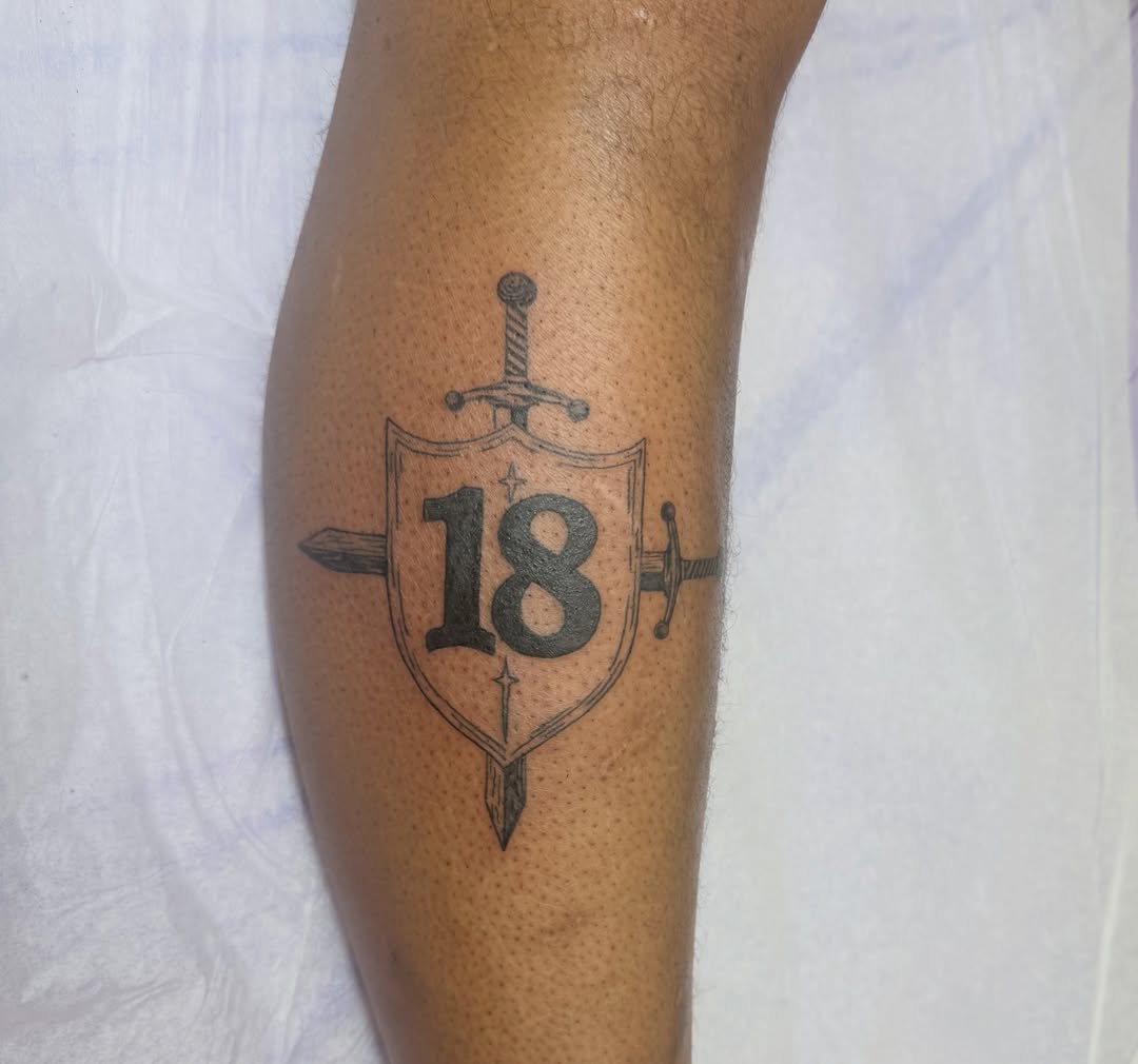 Shield with sword and number 18 black and grey tattoo on the forearm by Ink About Me - Amanda’s Fine Lines