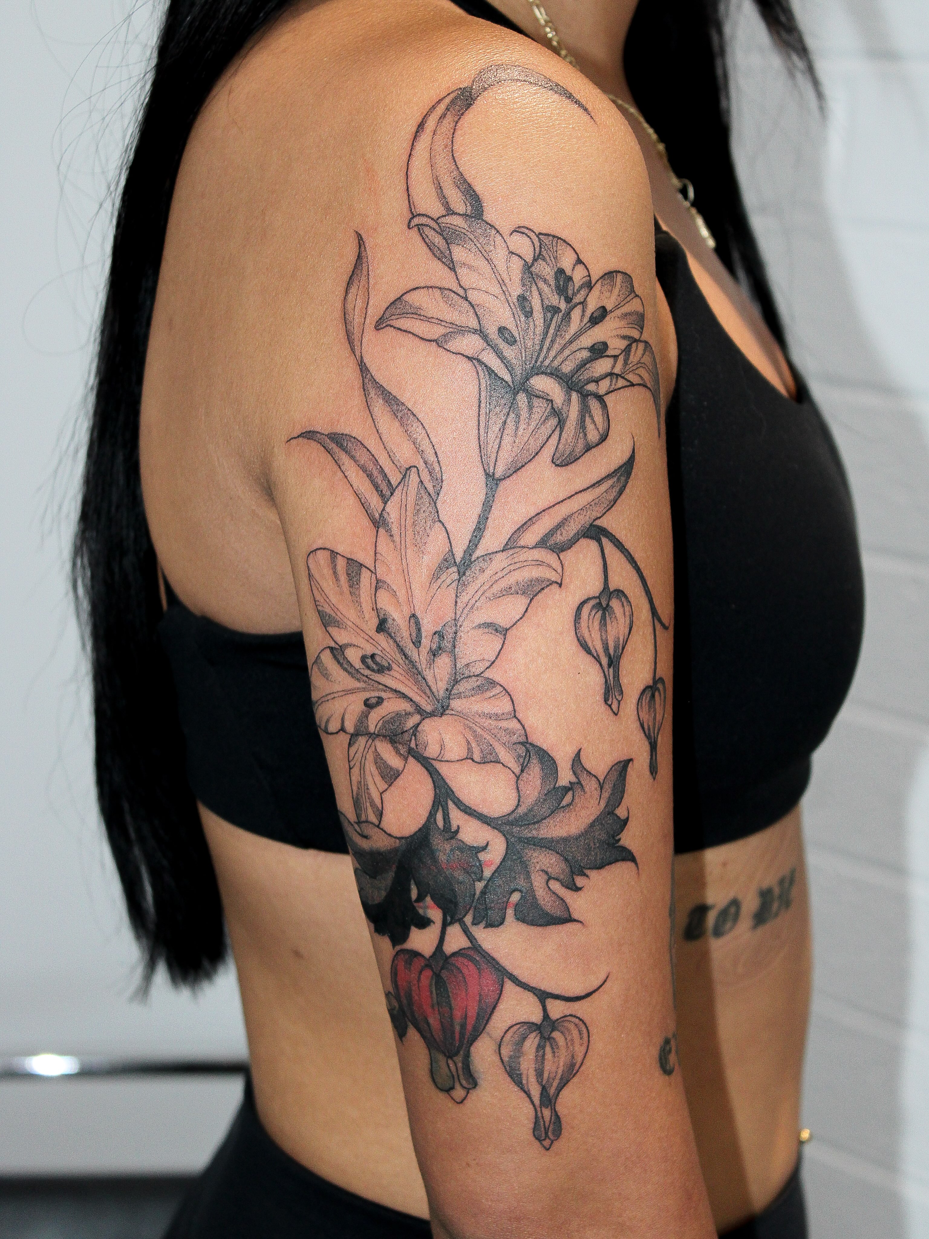 Lilies and heart-shaped flowers black and grey tattoo on the upper arm by Julianna
