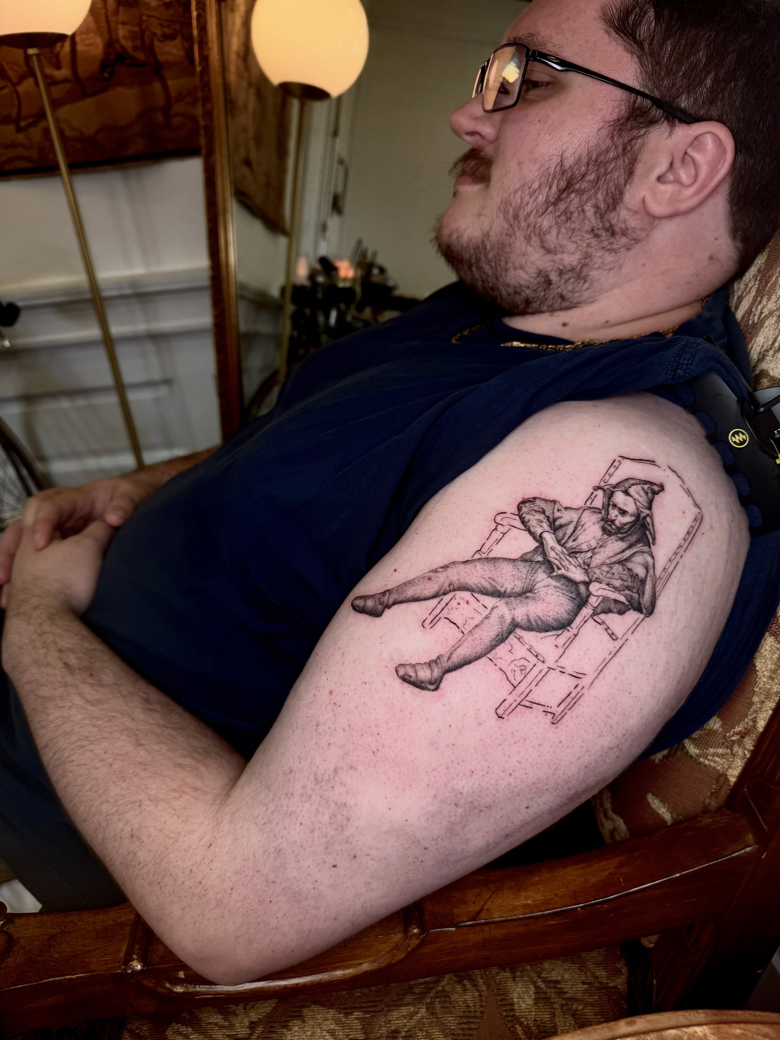Man in chair realism tattoo on the upper arm by Ian Saner